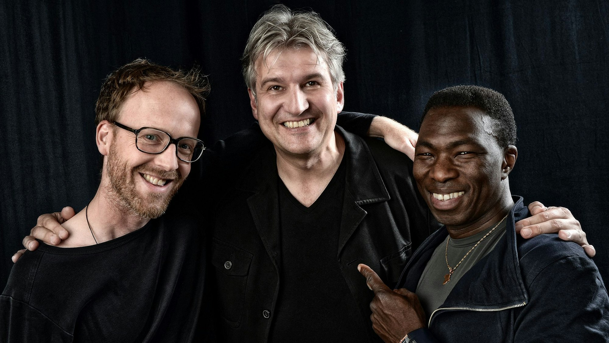 Drei Männer stehen Arm in Arm nebeneinander und lächeln in die Kamera.