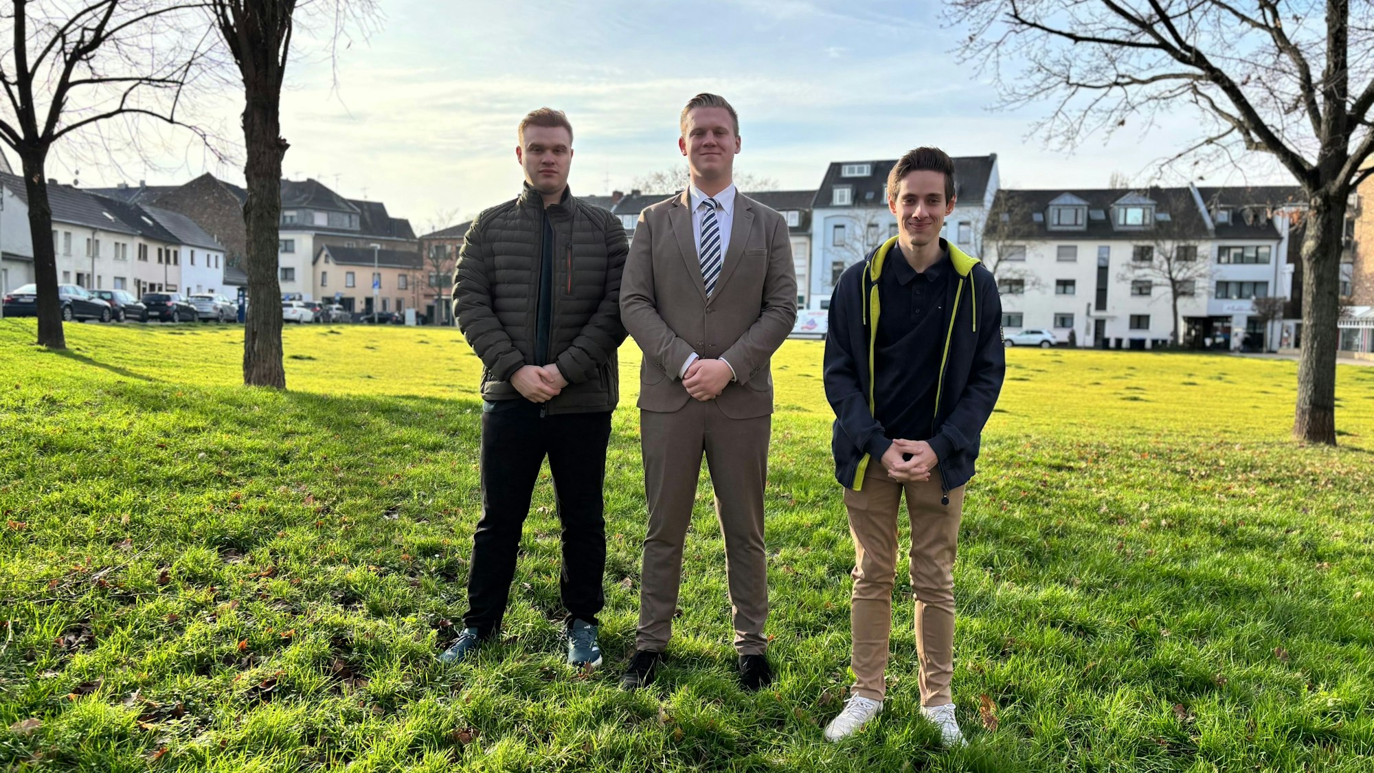 Die drei jungen Männer stehen nebeneinander auf einer Wiese, die Hände vor sich verschränkt.