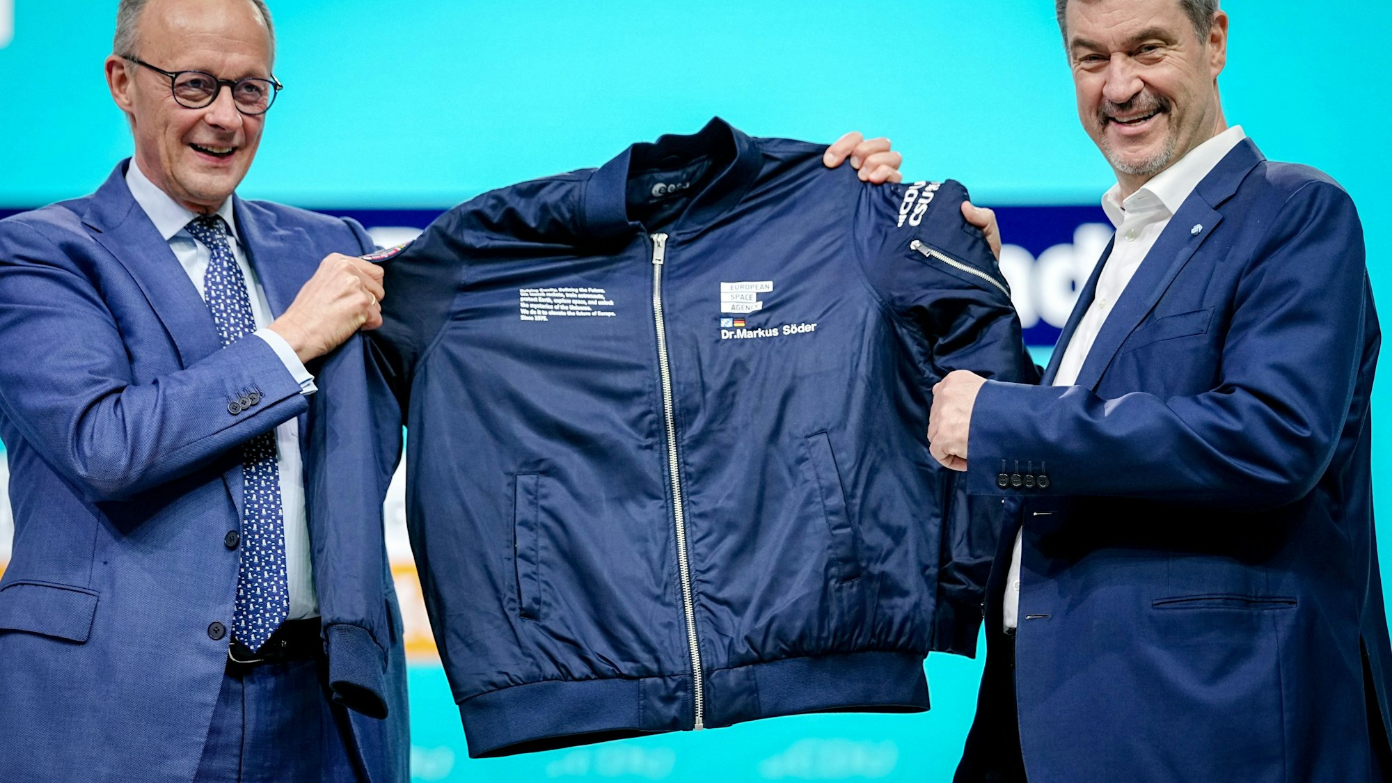 Friedrich Merz (l, CDU) überreicht Markus Söder, CSU-Vorsitzender und Ministerpräsident von Bayern, beim CDU-Bundesparteitag auf der Bühne eine Jacke mit der Aufschrift European Space Agency.