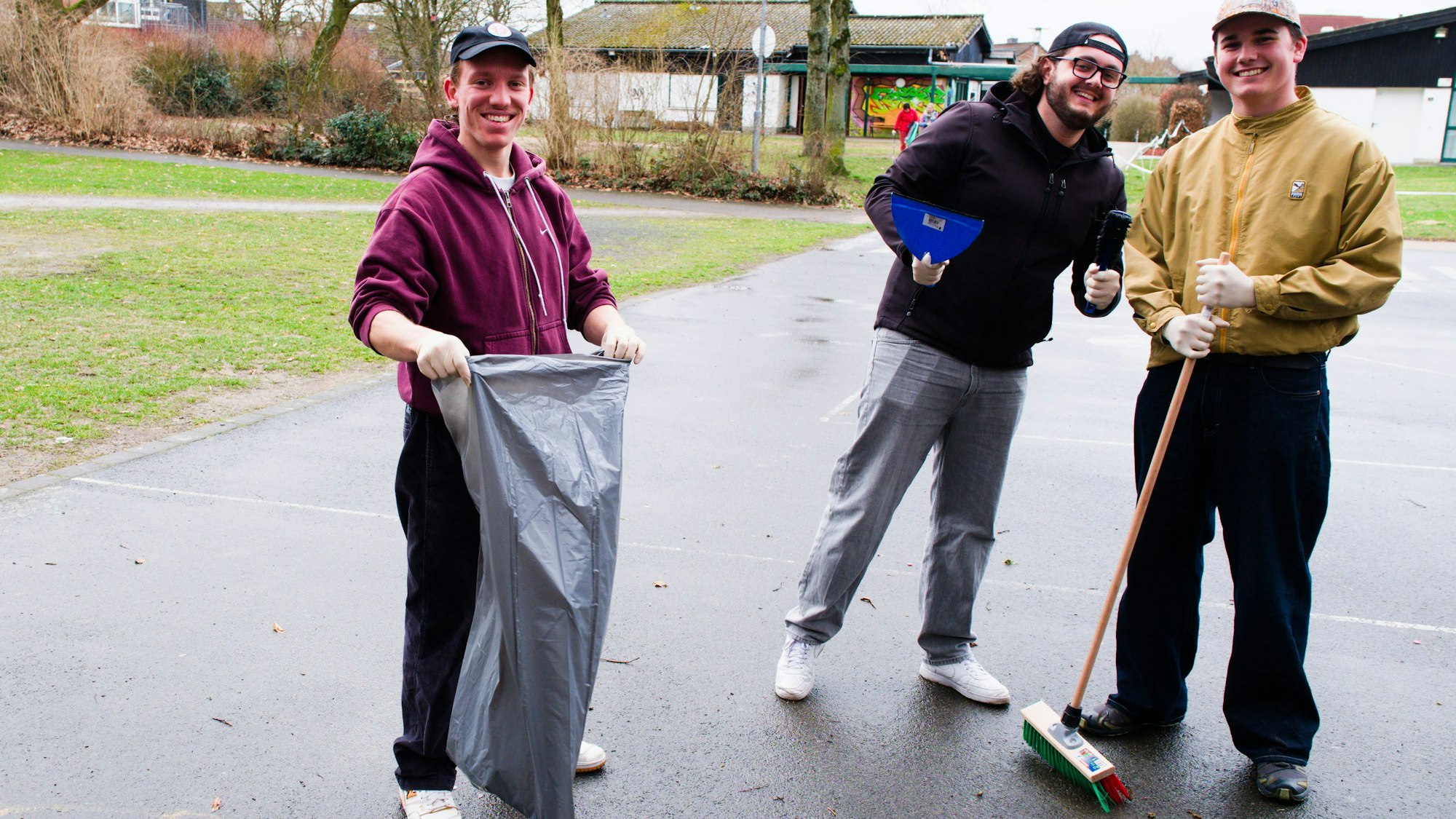 Drei junge Männer mit einem grauen Müllsack, Handfeger und Schaufel und einem Besen, lächelnd