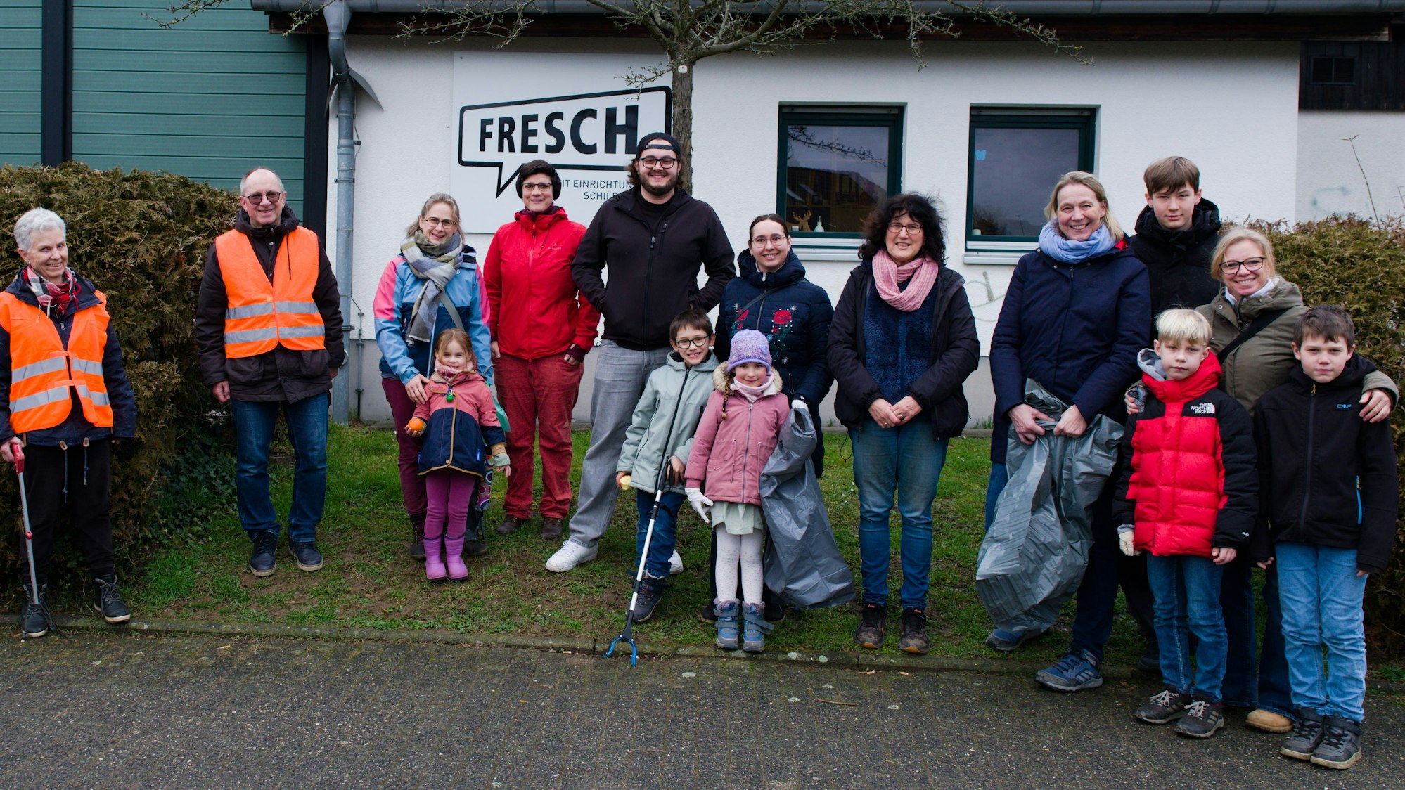 Eine Gruppe Alt und Jung steht, mit Müllsammel-Werkzeug ausgerüstet, vor einem Gebäude mit der Aufschrift Fresch