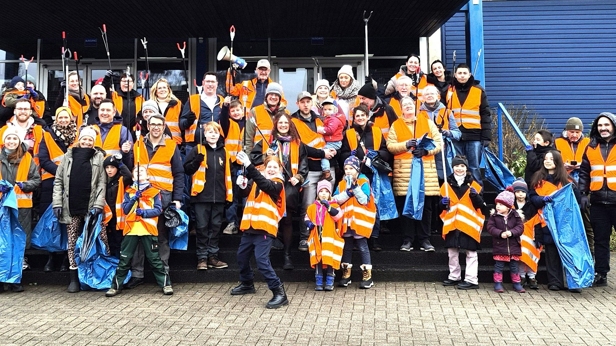 Eine große Gruppe Menschen mit orangen Warnwesten halten blaue Müllsäcke und Greifer in den Händen
