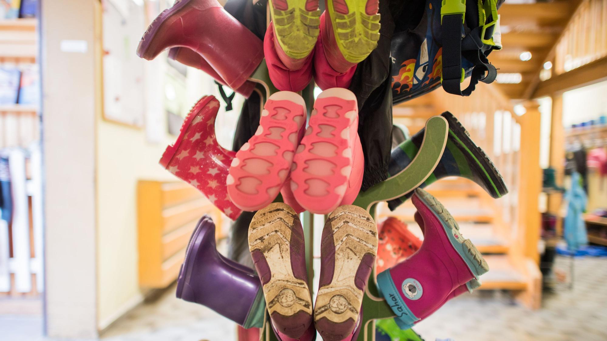 Bunte Kinder-Gummistiefel stecken paarweise auf einem hölzernen Ständer in einem Kindergarten-Raum