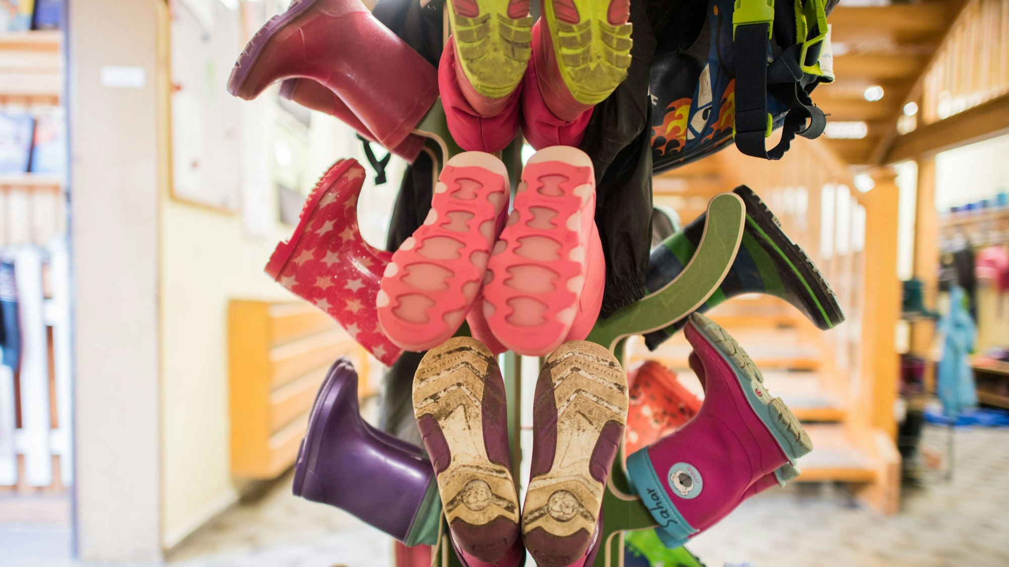Bunte Kinder-Gummistiefel stecken paarweise auf einem hölzernen Ständer in einem Kindergarten-Raum