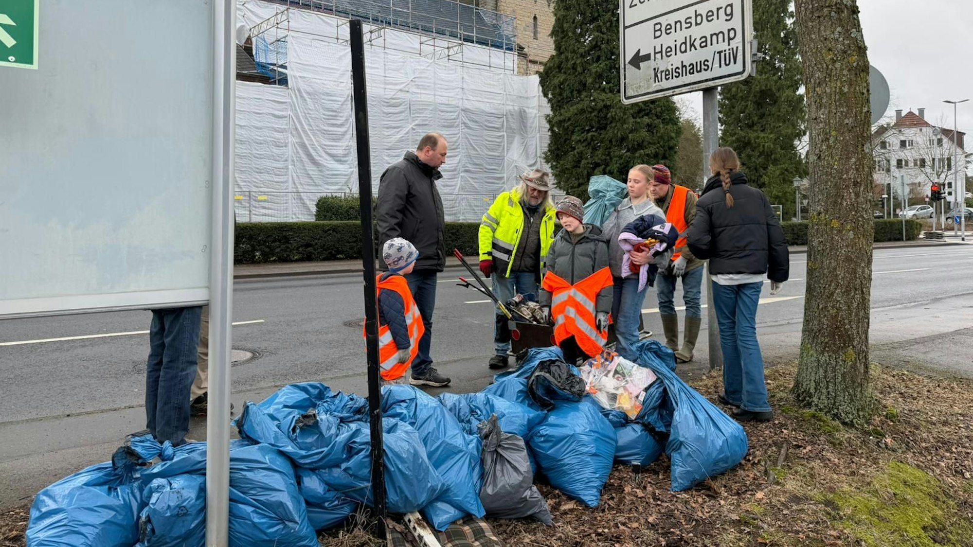 eine große Zahl blauer gefüllter Müllsäcke an einer Straße, daneben Menschen mit Sammel-Werkzeug und weiteren vollen Tüten in den Händen
