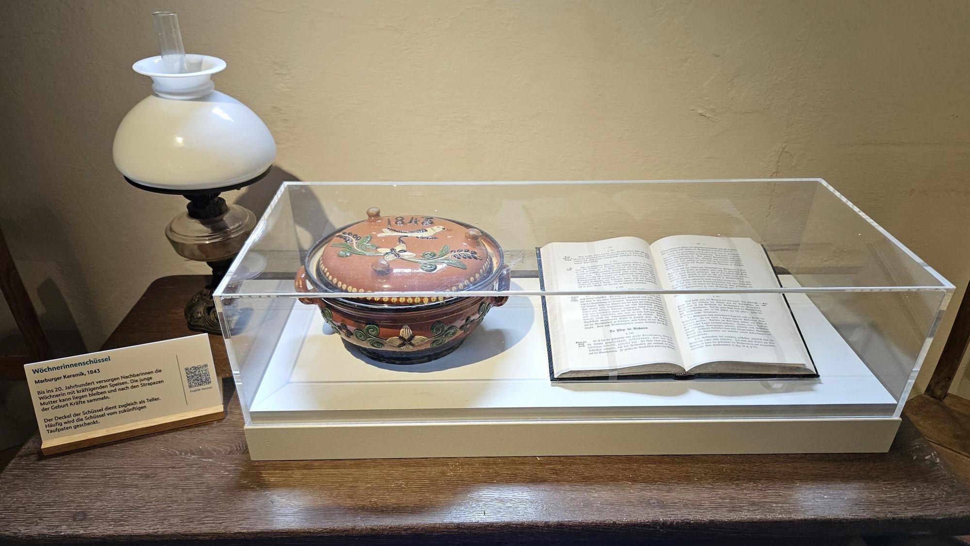 Blick auf eine Vitrine mit einer Wöchnerinnenschüssel und einem aufgeschlagenen Buch.