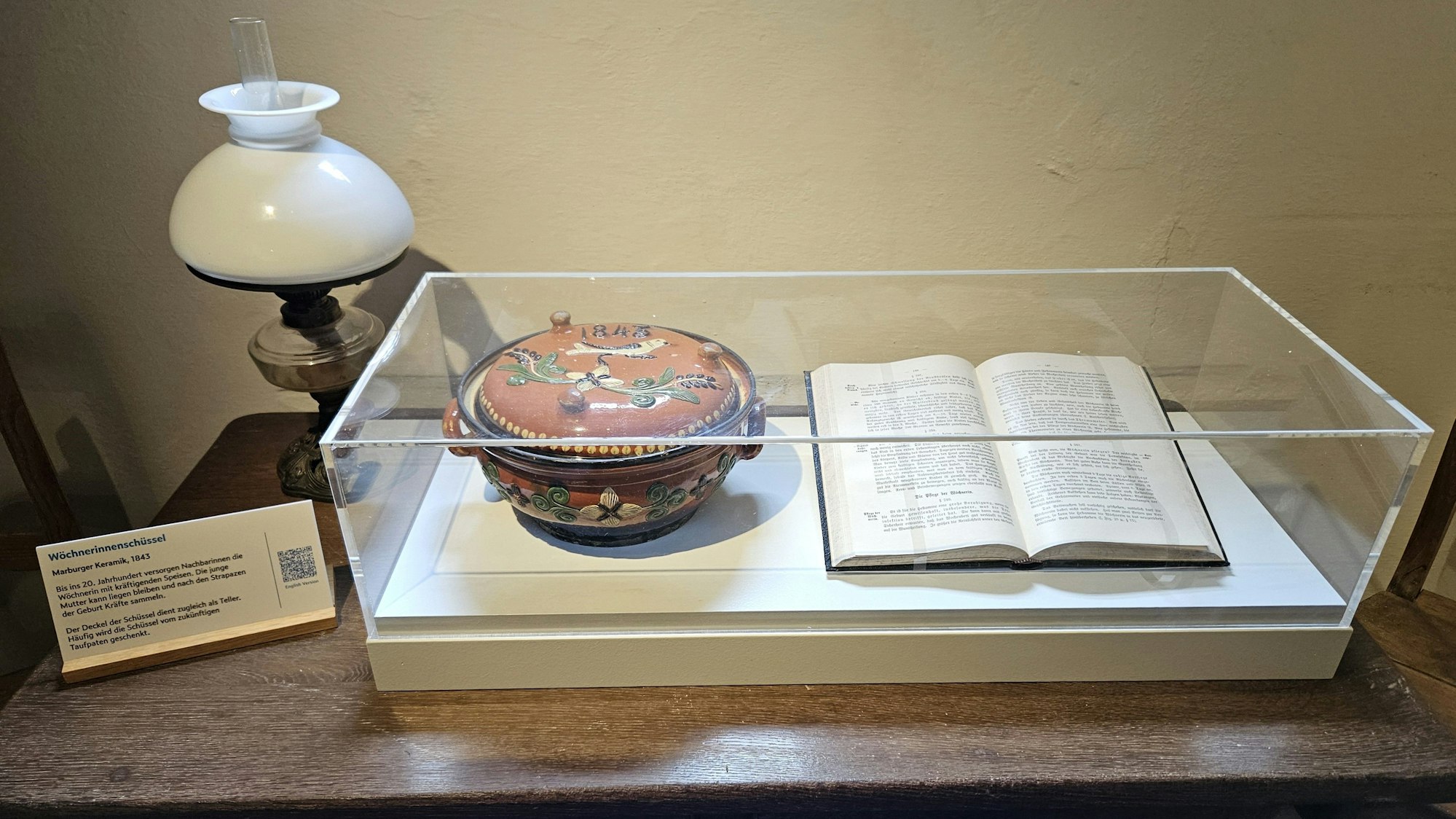 Blick auf eine Vitrine mit einer Wöchnerinnenschüssel und einem aufgeschlagenen Buch.
