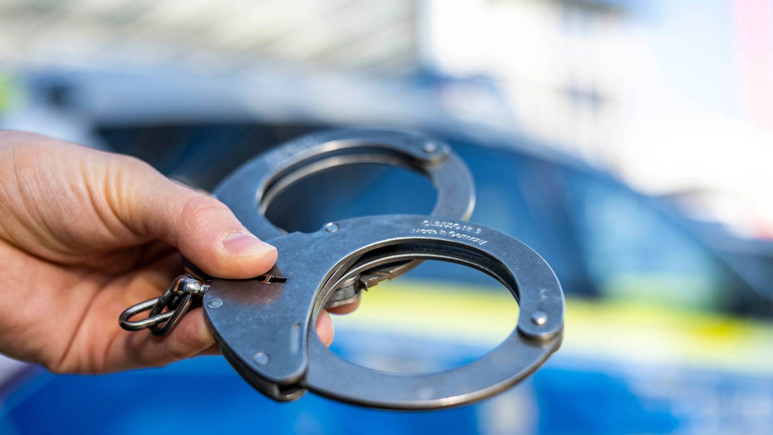Ein Beamter hält Handschellen vor einen Streifenwagen der NRW-Polizei.