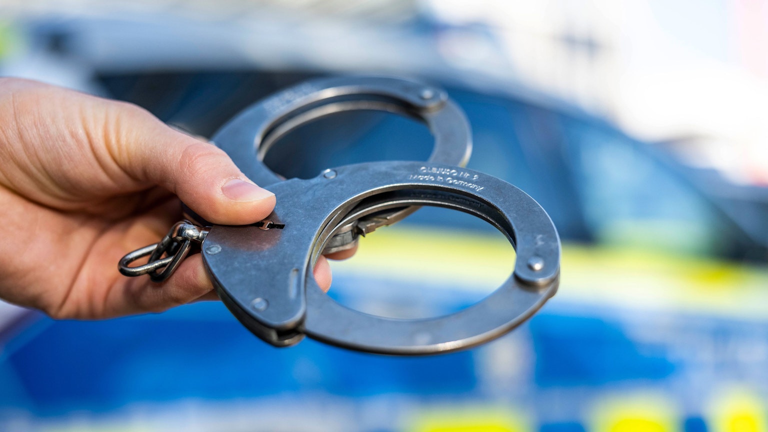 Ein Beamter hält Handschellen vor einen Streifenwagen der NRW-Polizei.