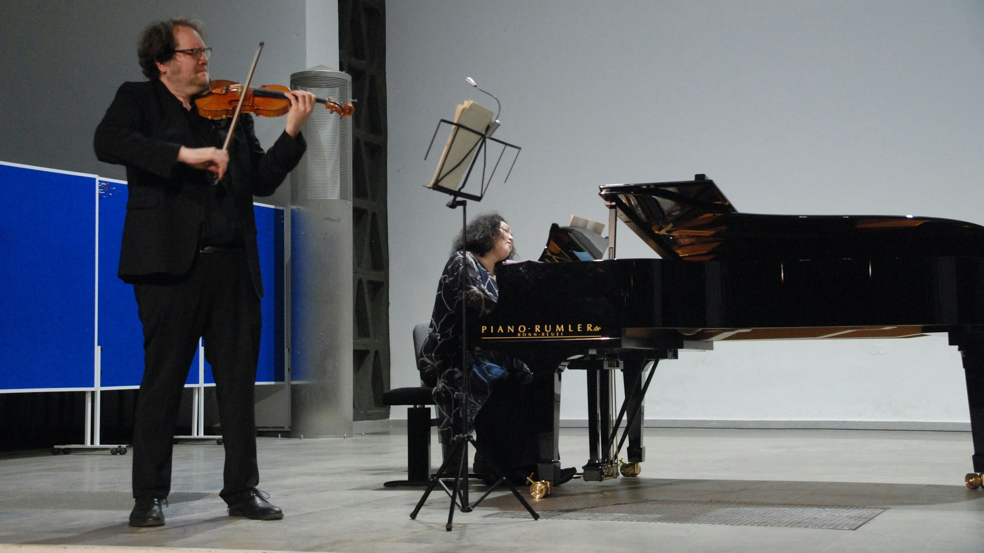 Im Vordergrund links steht ein Mann, der Geige spielt. Rechts sitzt eine Frau am Flügel.