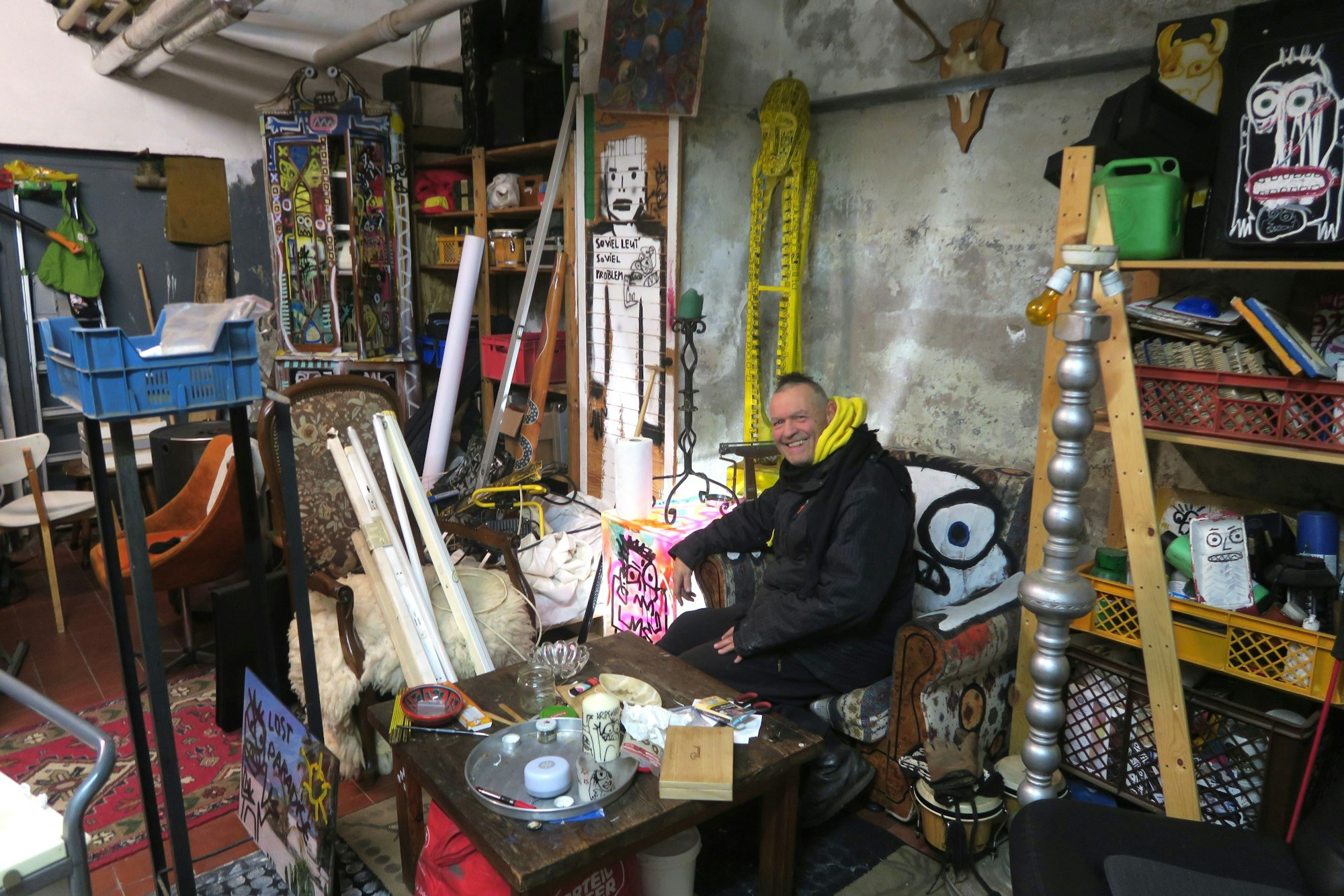 Ein Man sitzt auf einem bunten Sessel, um ihn herum Krempel auf Tischen und in Regalen.