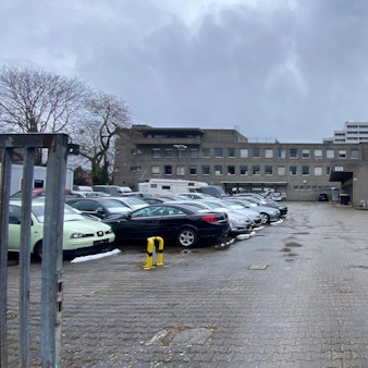 Zu sehen sind eine als Parkplatz genutzte Fläche sowie im Hintergrund Gebäude.