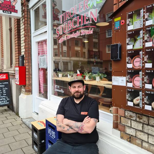 Ein Mann mit Bart und Basecap sitzt mit verschränkten Armen vor einem Café auf einem Hocker.