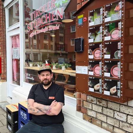 Ein Mann mit Bart und Basecap sitzt mit verschränkten Armen vor einem Café auf einem Hocker.