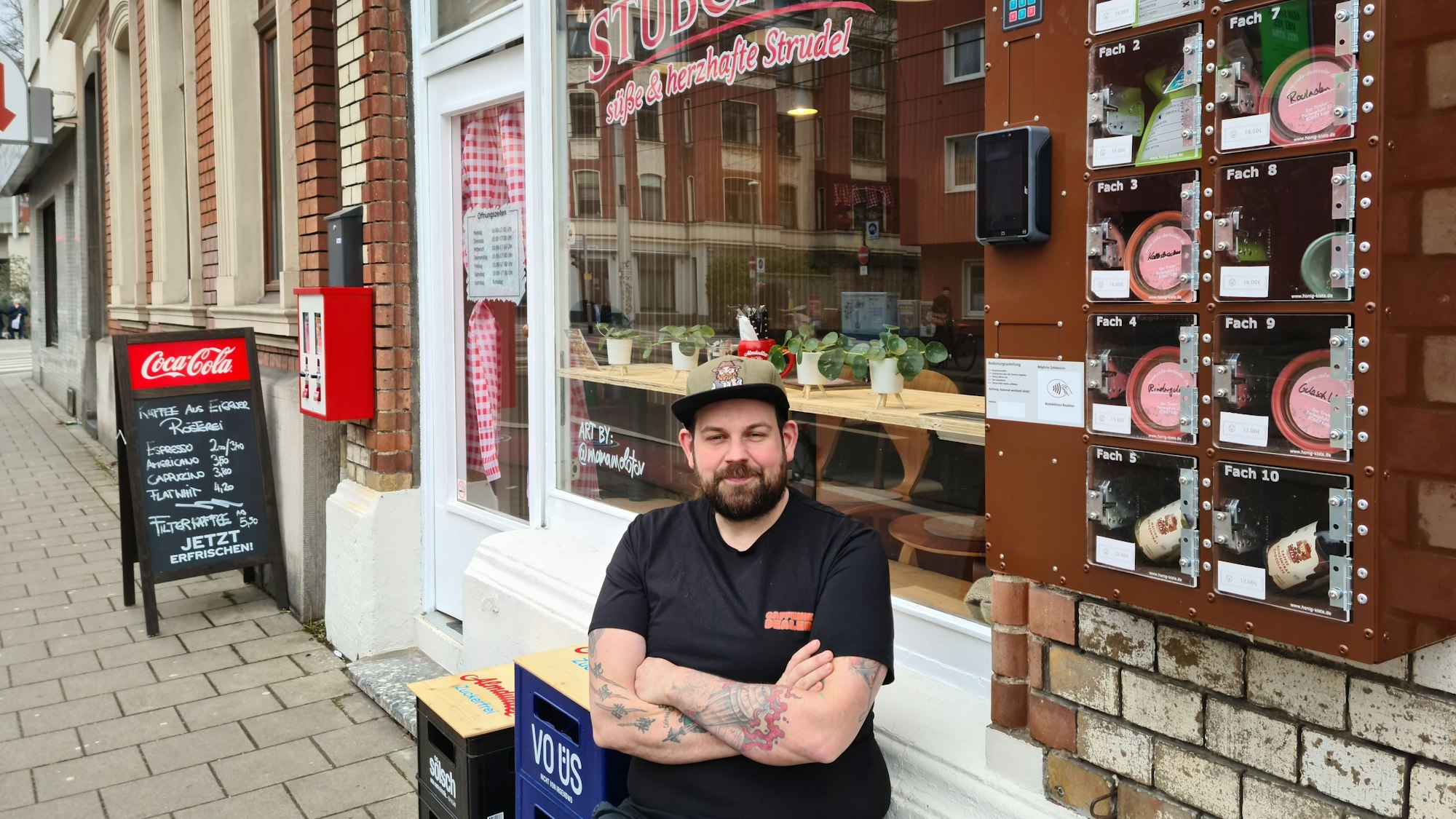 Ein Mann mit Bart und Basecap sitzt mit verschränkten Armen vor einem Café auf einem Hocker.
