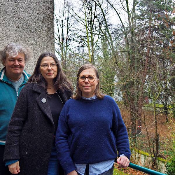 Ein Mann und zwei Frauen stehen auf einem Balkon, im Hintergrund sind Bäume in einem großen Hinterhof-Grundstück zu sehen.