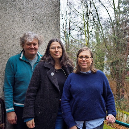 Ein Mann und zwei Frauen stehen auf einem Balkon, im Hintergrund sind Bäume in einem großen Hinterhof-Grundstück zu sehen.