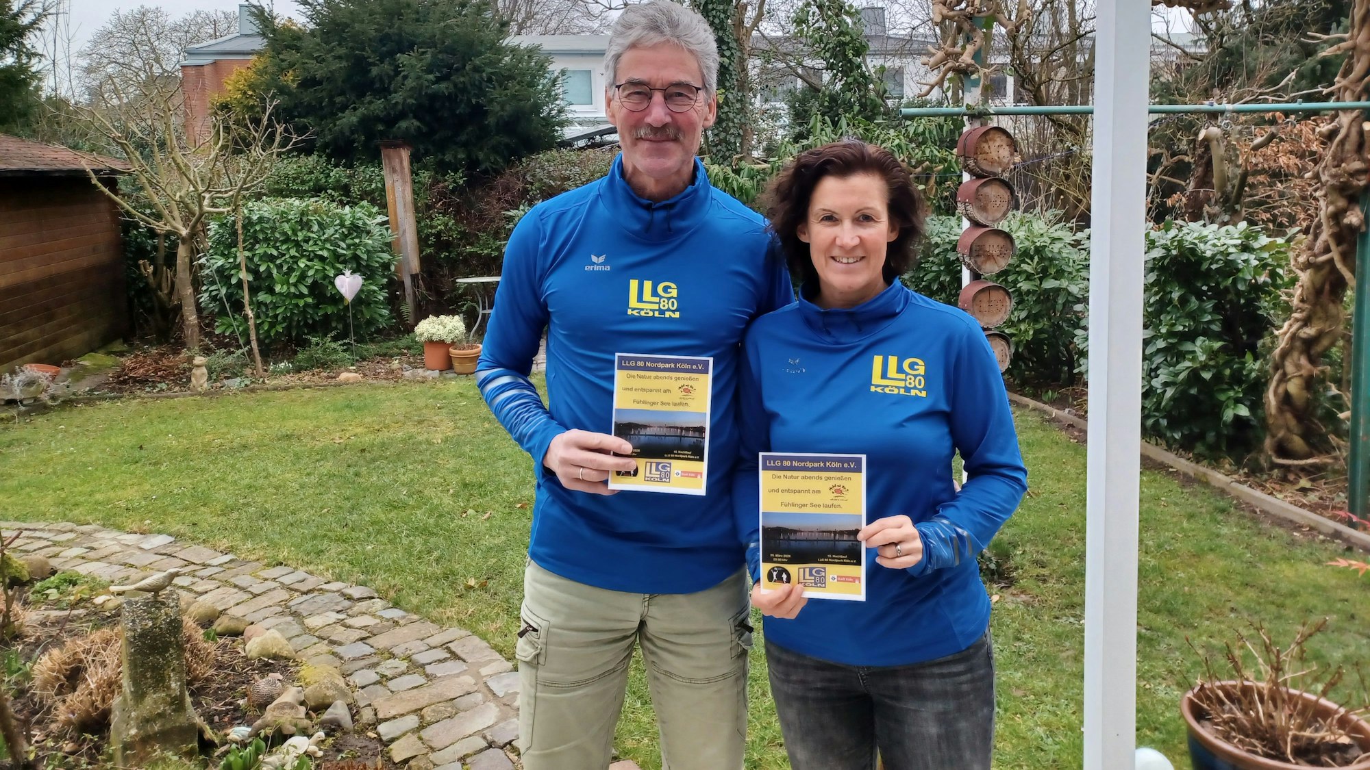 Ein Mann und eine Frau stehen nebeneinander in einem Garten. Sie halten Flyer zum Nachtlauf in der Hand.