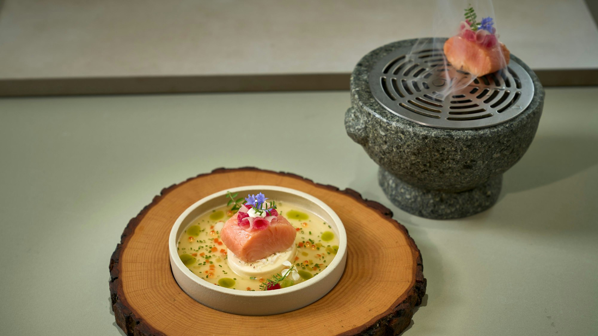 Eine Holzscheibe, darauf ein kleiner Teller mit einem Stückchen Lachs und ein Gefäß, aus dem Dampf aufsteigt, ebenfalls mit einem verzierten Stückchen Lachs.