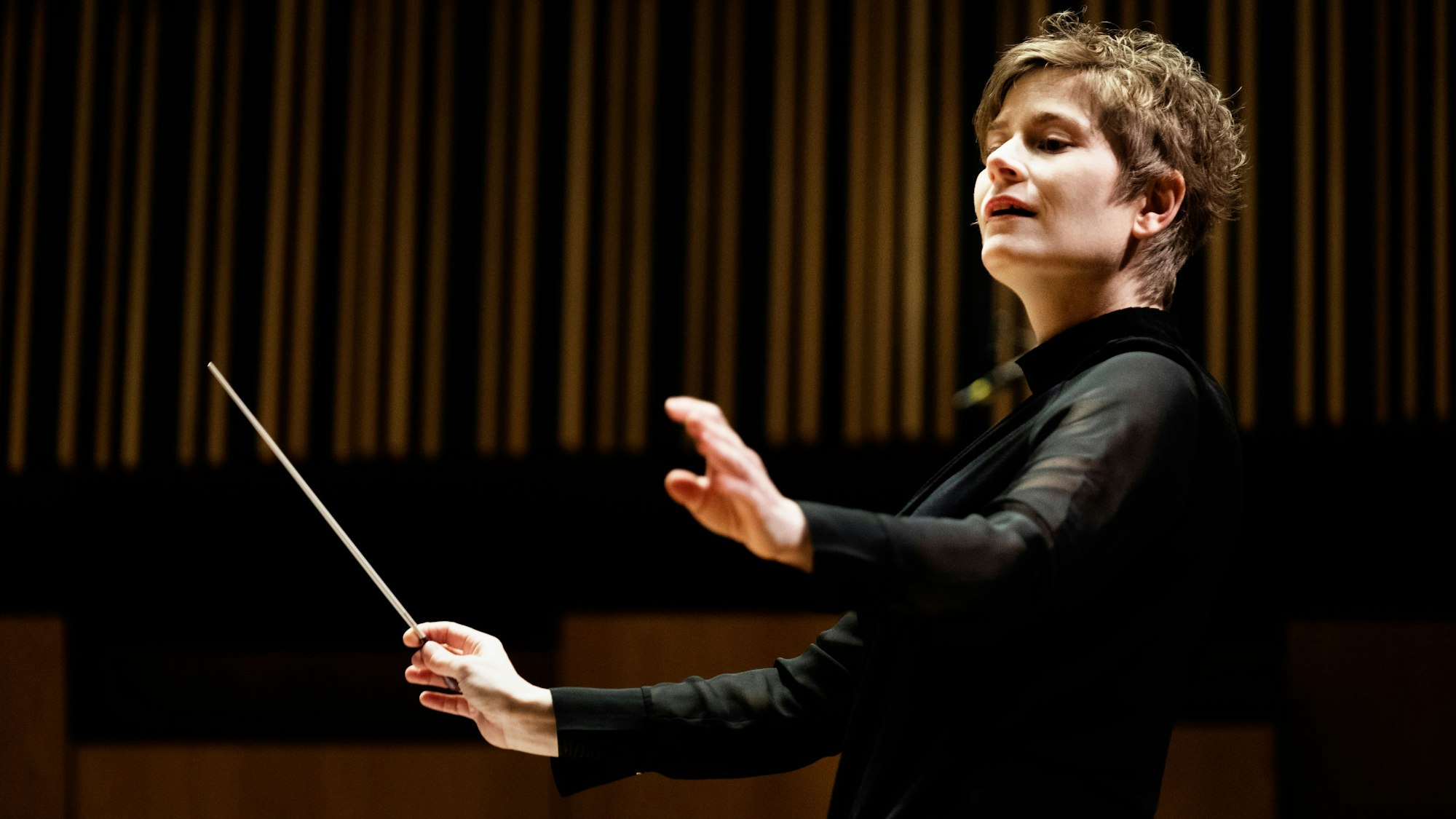 Eine Dirigentin hält in ihrer rechten Hand einen Taktstock und breitet die Arme aus.