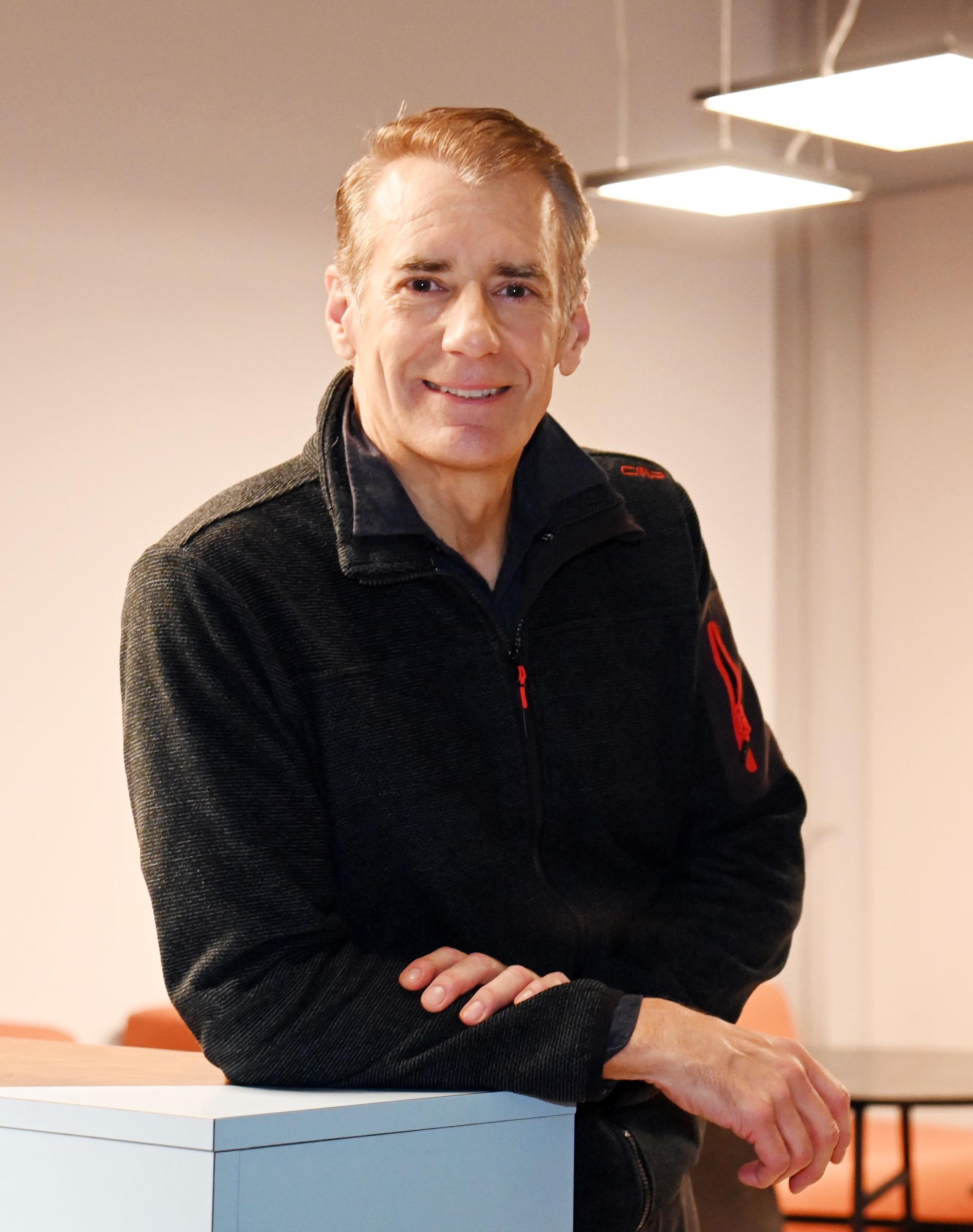 Ein Mann mit grauem Haar und schwarzem Pullover steht an einem Tisch.