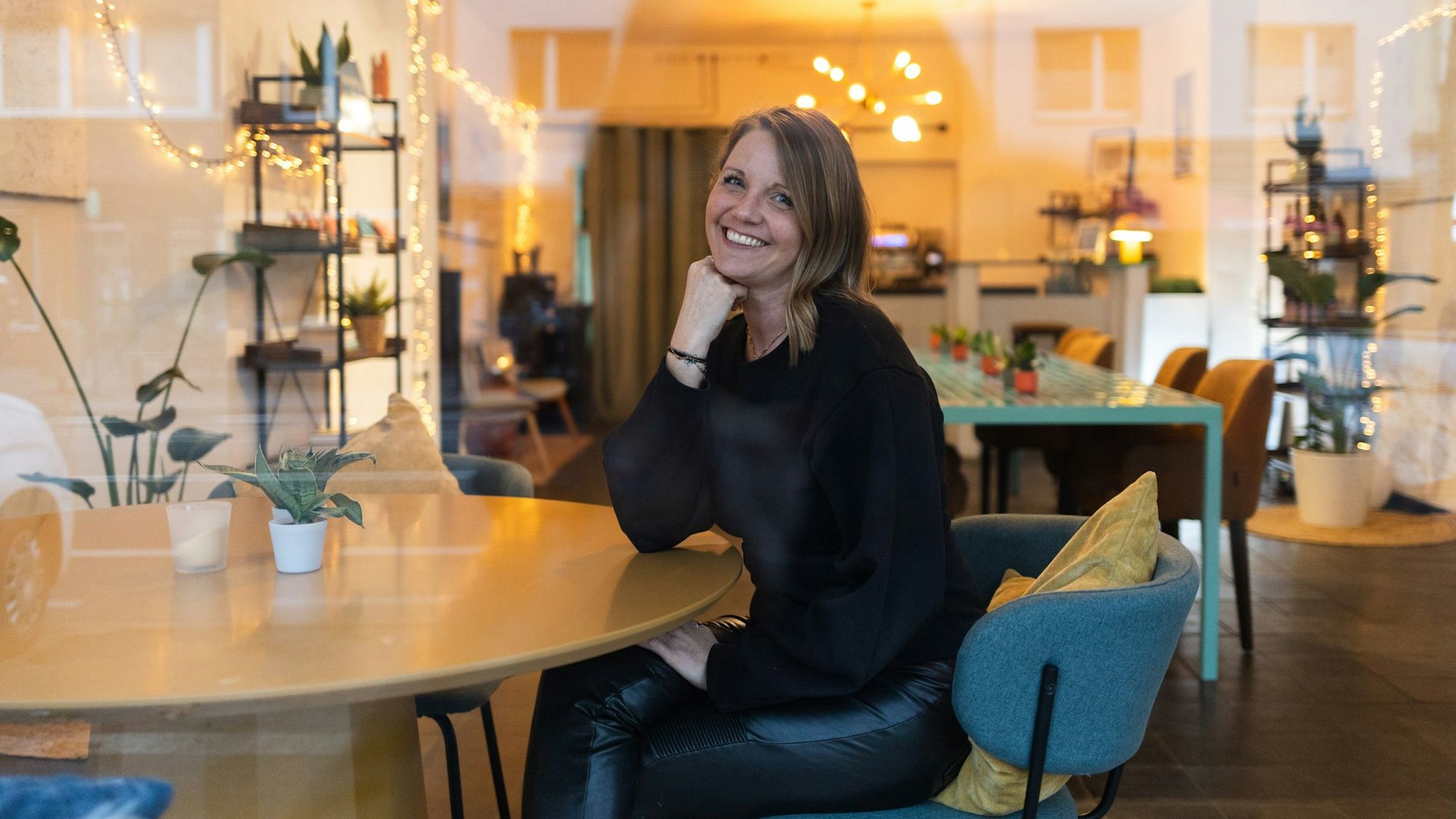 Eine Frau sitzt auf einem Stuhl und lächelt in die Kamera. Hinter ihr sieht man einen Raum mit Tischen und einer Bar.