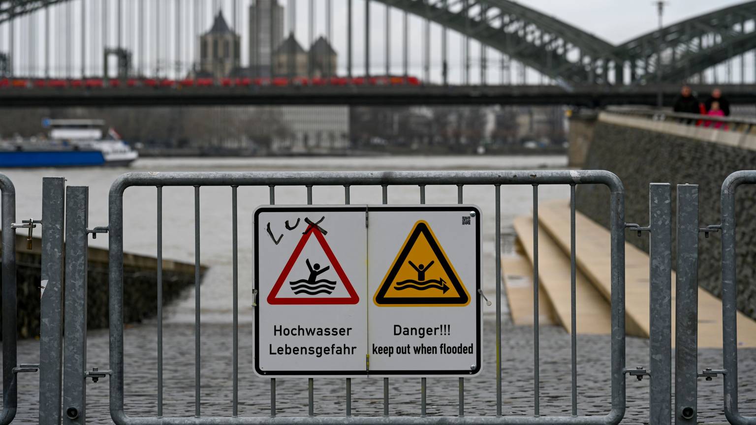 Der Rheinboulevard in Köln ist zum Teil überschwemmt. Im Hintergrund ist die Hohenzollernbrücke zu sehen. Ein Schild warnt vor Hochwasser.