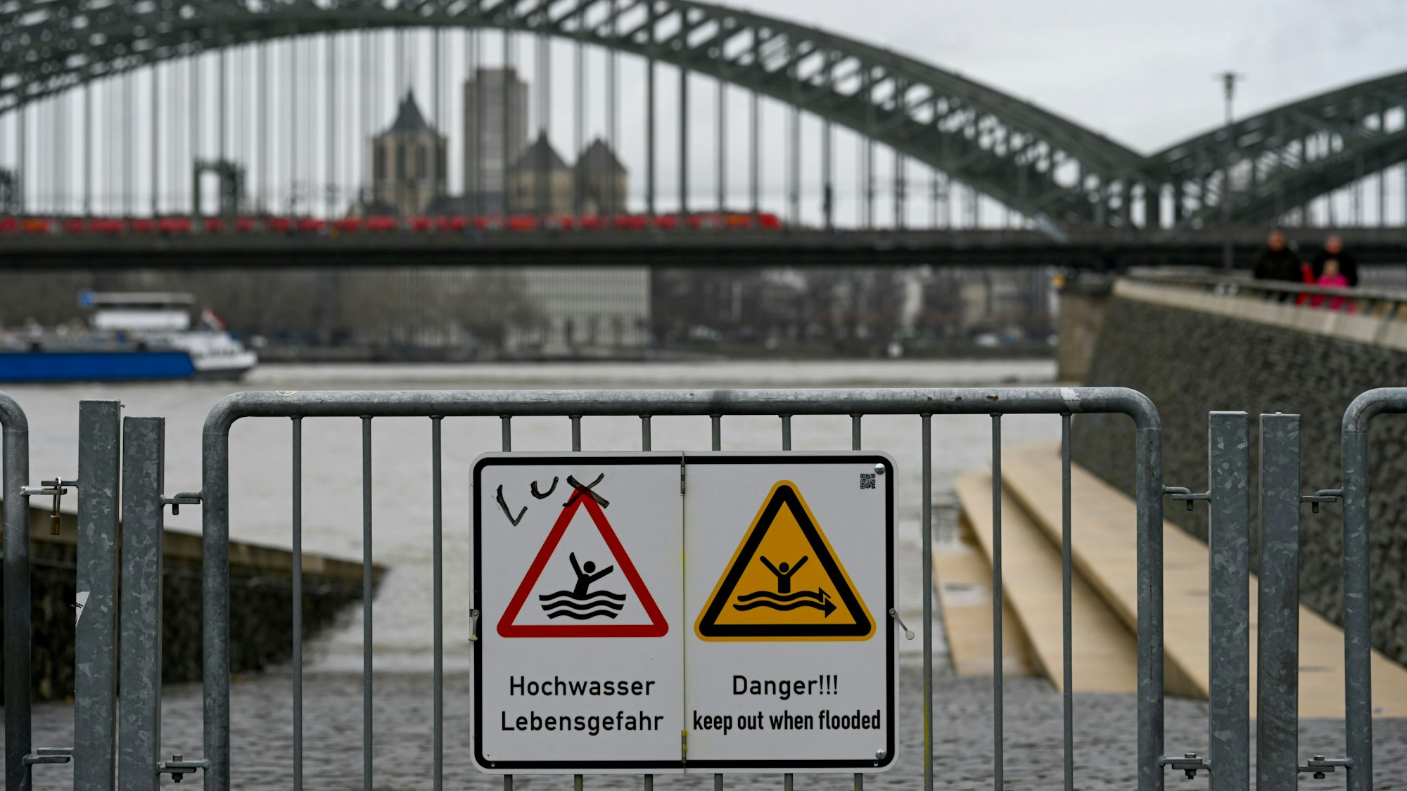 Der Rheinboulevard in Köln ist zum Teil überschwemmt. Im Hintergrund ist die Hohenzollernbrücke zu sehen. Ein Schild warnt vor Hochwasser.