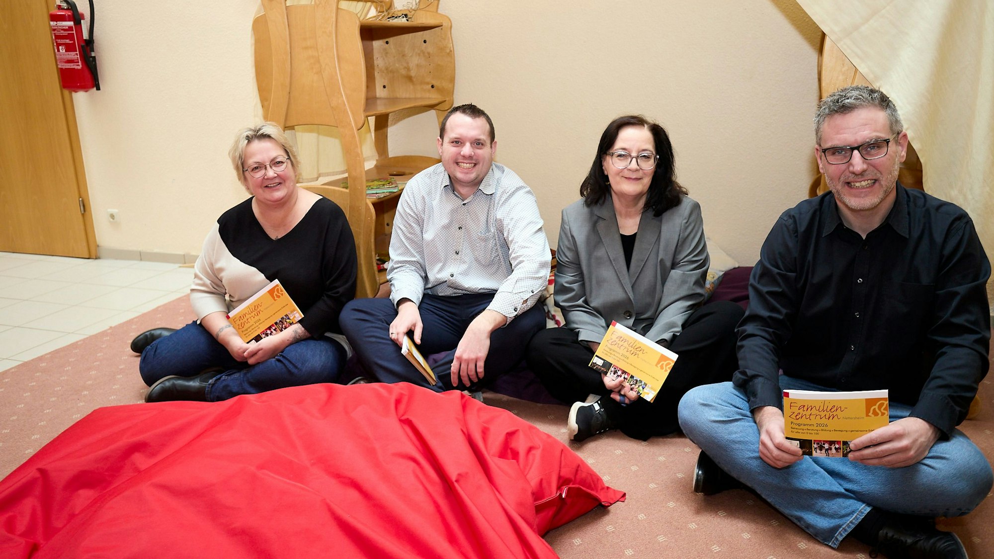 Die genannten Personen sitzen im Schneidersitz auf dem Boden, vor ihnen kueger ein rotes Riesenkissen. Alle halten das Programmheft in den den Händen.