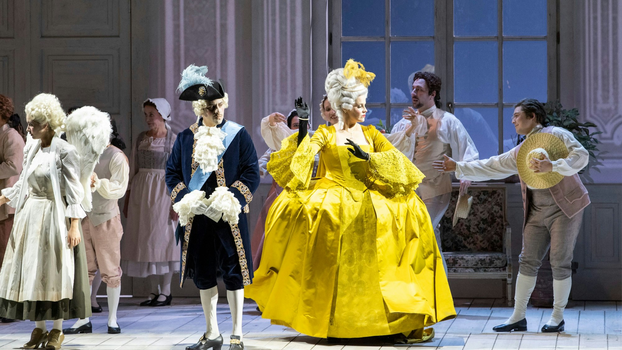 Eine Frau steht auf einer Bühne und trägt ein gelbes Kleid. Um sie herum stehen andere verkleidete Sängerinnen und Sänger.