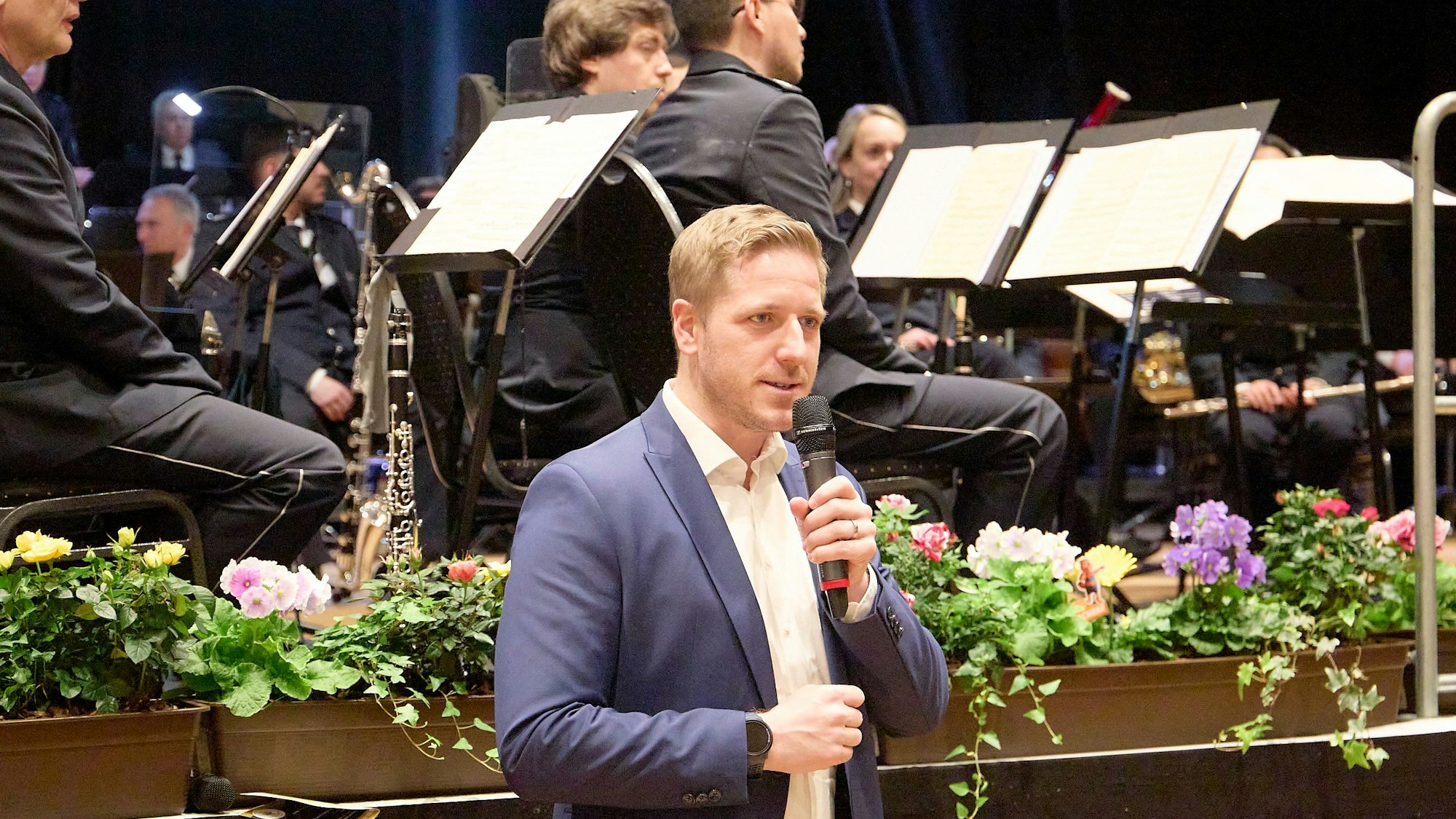 Landrat Markus Ramers sprich in ein Mikrofon. Hinter dem Politiker sitzen Mitglieder des NRW-Landespolizeiorchesters auf einer höher gelegenen Bühne.