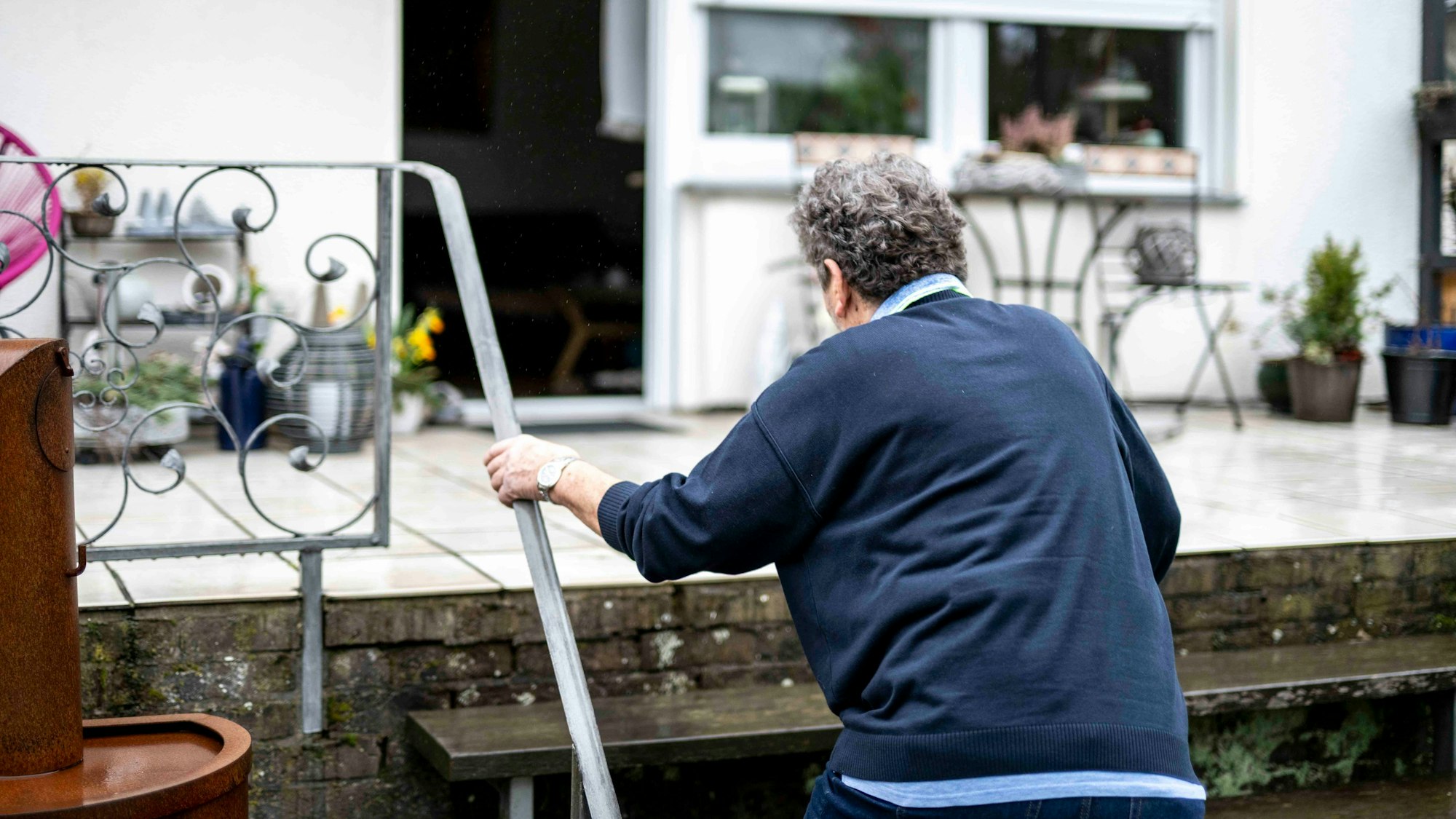 Ein älterer Mann ist von hinten zu sehen, wie er sich an einem Geländer festhält und eine Treppe zu einer Terrasse an einem Haus hochgeht.