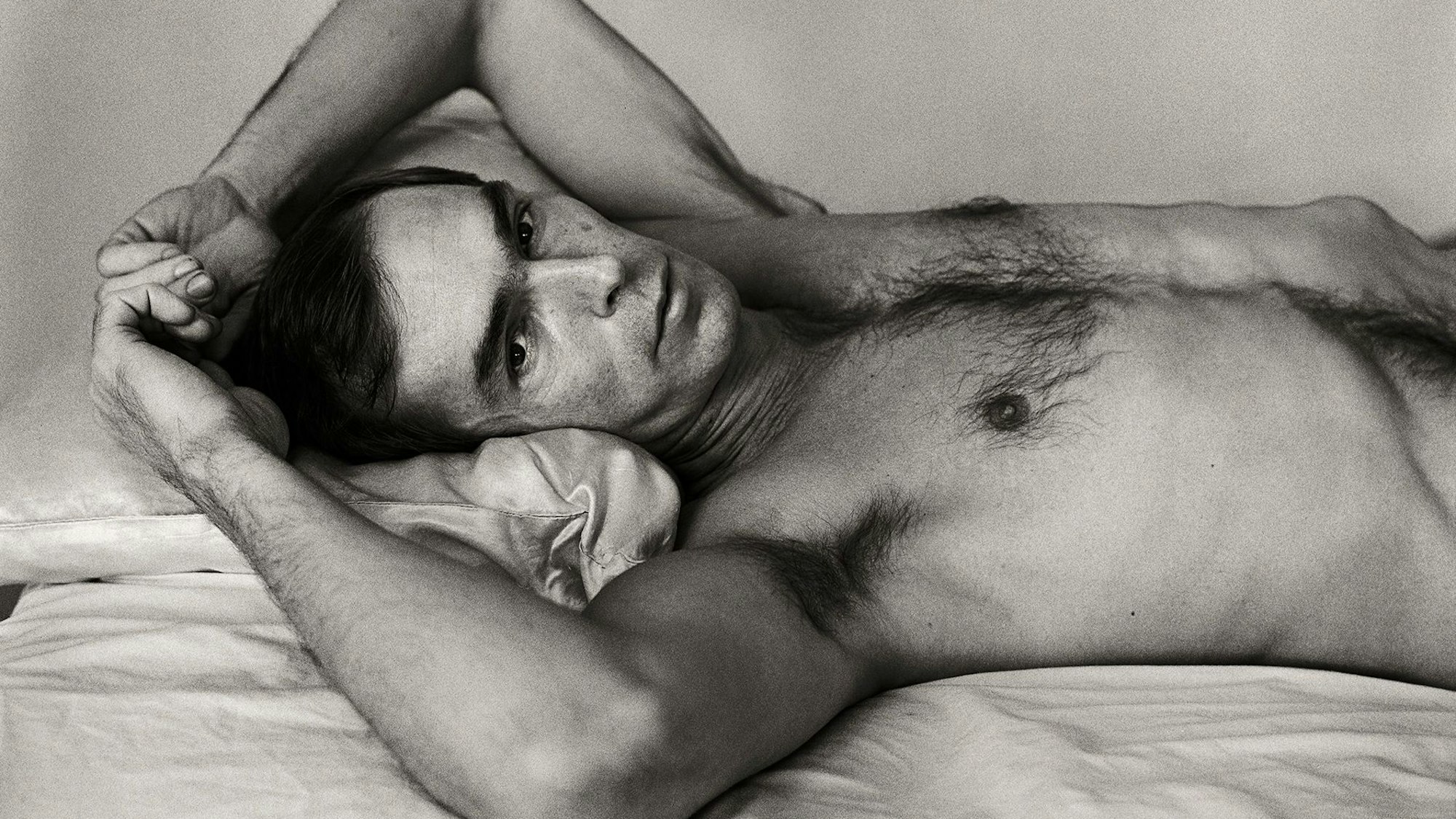 Peter Hujar liegt auf einem Bett und schaut in die Kamera.