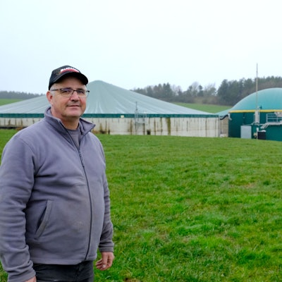 Landwirt Helmut Dahmen aus Lorbach steht auf einer Wiese, im Hintergrund ist eine Biogasanlage zu sehen.