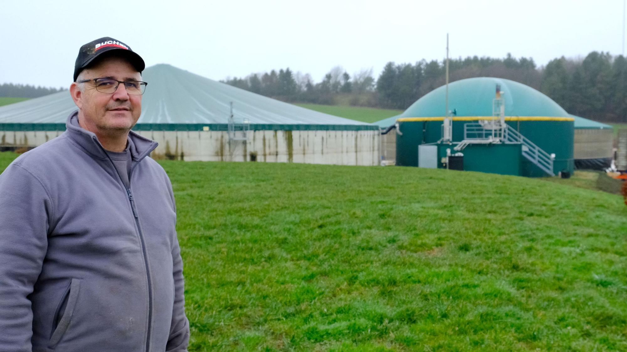Landwirt Helmut Dahmen aus Lorbach steht auf einer Wiese, im Hintergrund ist eine Biogasanlage zu sehen.