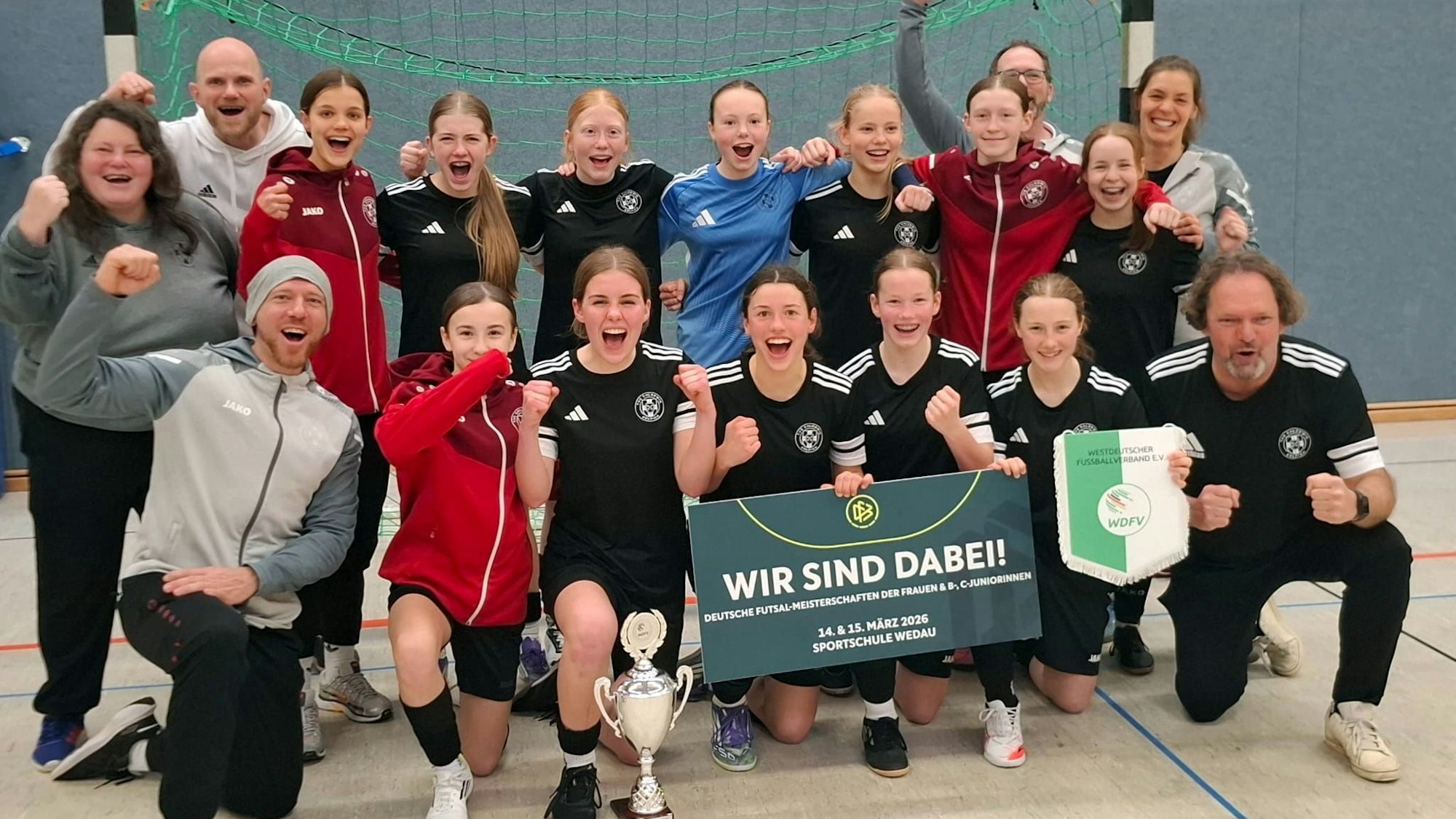 Spielerinnen und Betreuer jubeln. Sie hocken beziehungsweise stehen in zwei Reihen vor einem Tor.