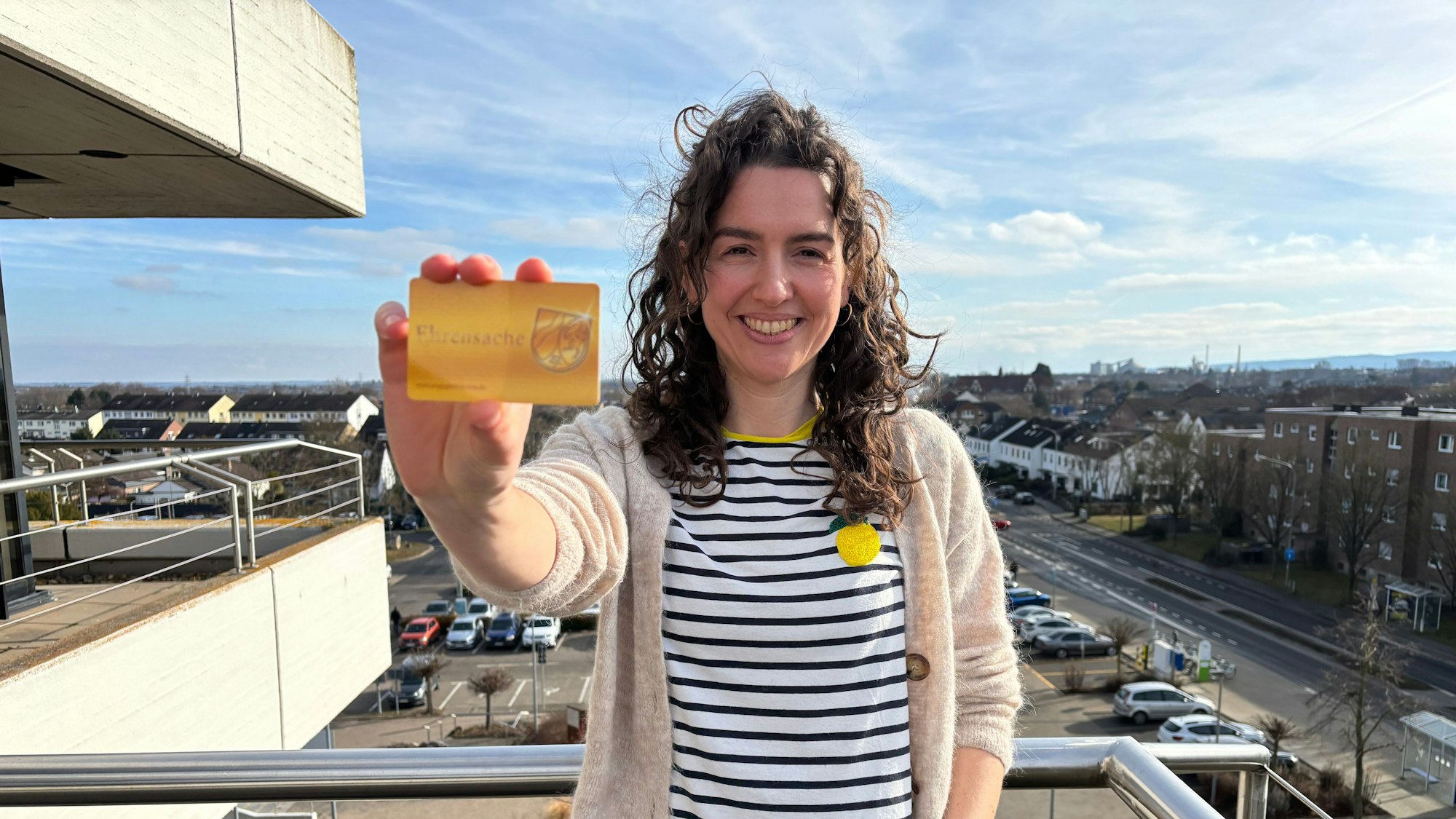 Vera Ahlbach steht lächelnd auf einem Balkon und hält eine Ehrenamtskarte in Richtung der Kamera.