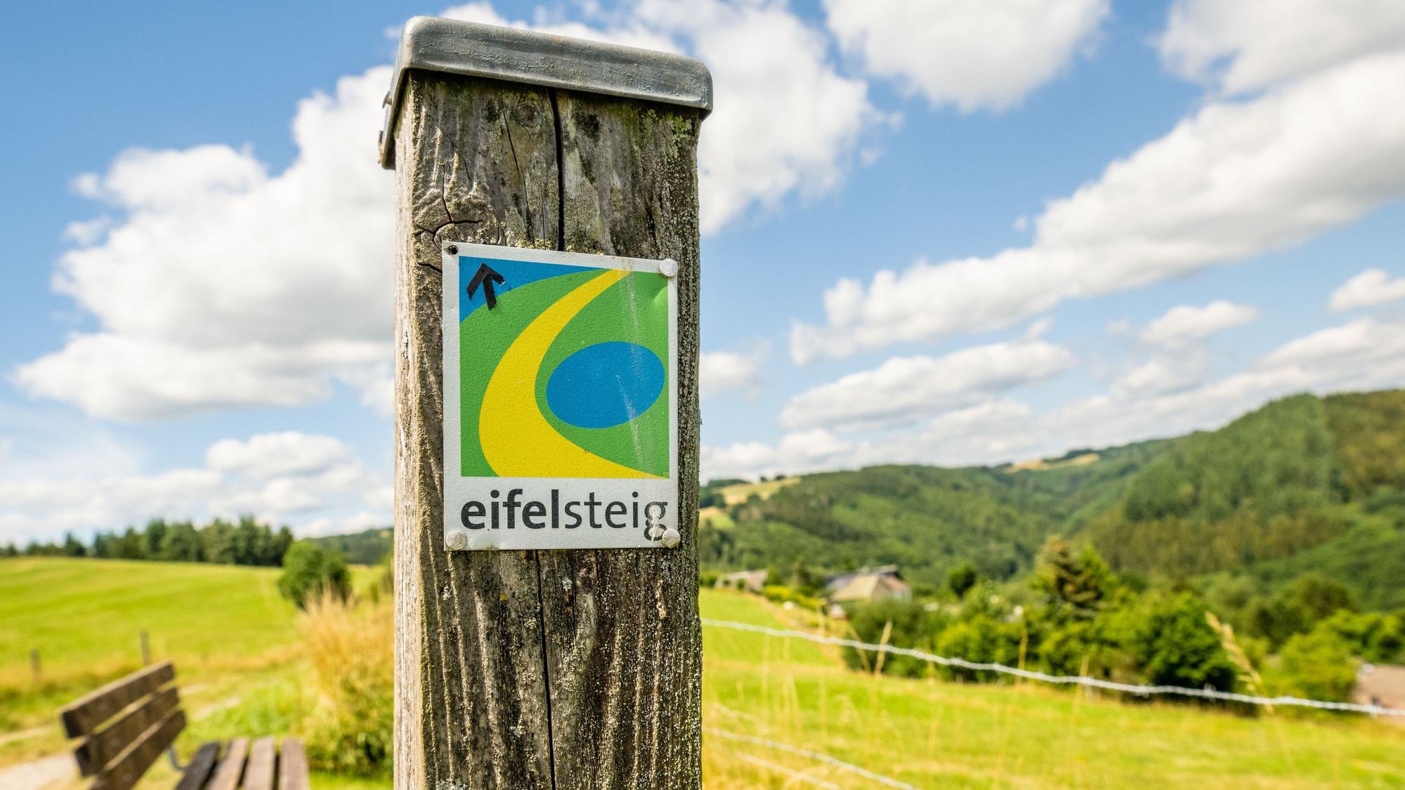 An einem Pfosten ist das Logo des Eifelsteigs befestigt. Im Hintergrund erstreckt sich eine grüne Hügellandschaft,