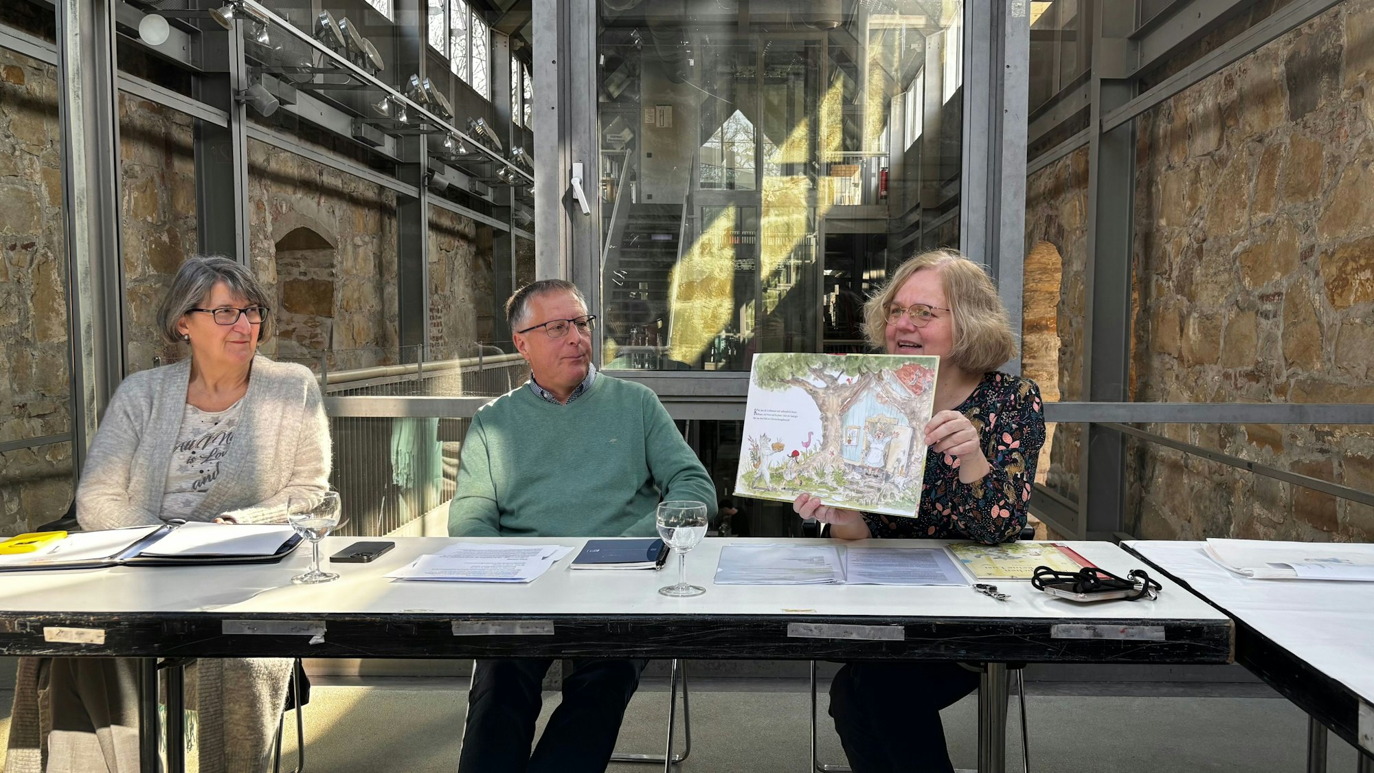 Zwei Frauen und ein Mann sitzen am Tisch. Die Leiterin hält das Bilderbuch in den Händen und erzählt.