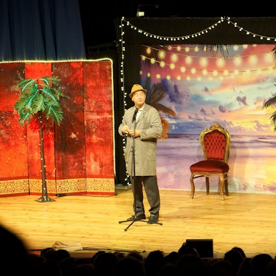 Gerhardt steht im Hausmeisterkittel und mit Hut auf der Theaterbühne.