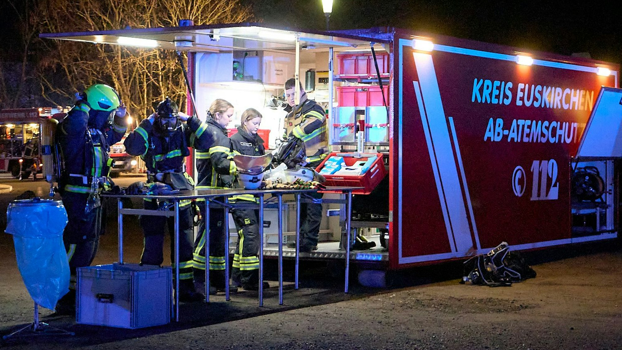 Vier Feuerwehrleute wechseln am Atemschutzcontainer des Kreises Euskirchen ihre Ausrüstung. Ein Mitarbeiter des Brandschutzzentrums unterstützt sie.