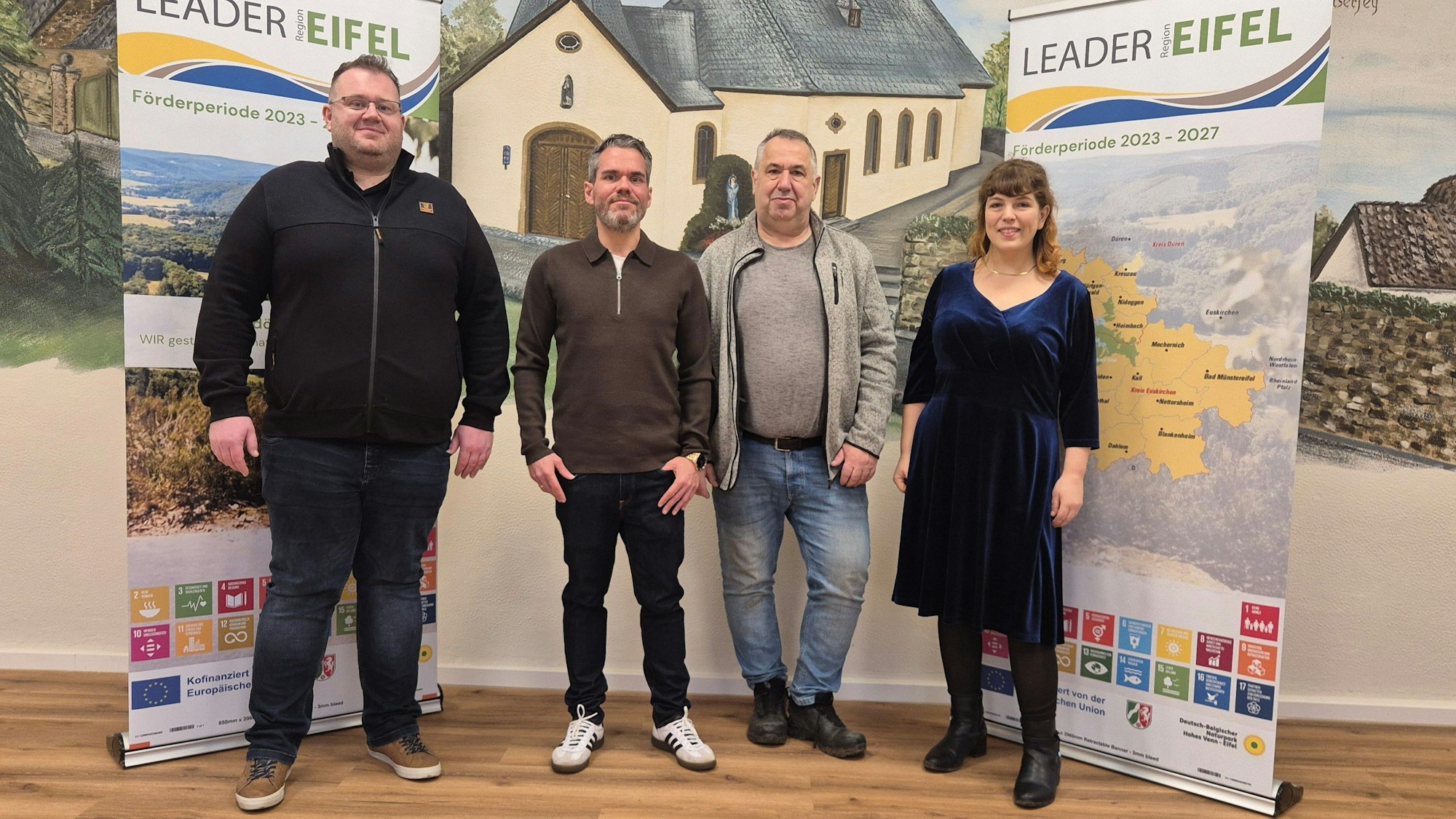 Drei Männer und eine Frau stehen vor einem Wandgemälde, rechts und links hängt Werbung für das Leader-Projekt.