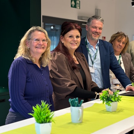 Die Obengenannten stehen versetzt an einem Stehtisch und lächeln in Richtung der Kamera.