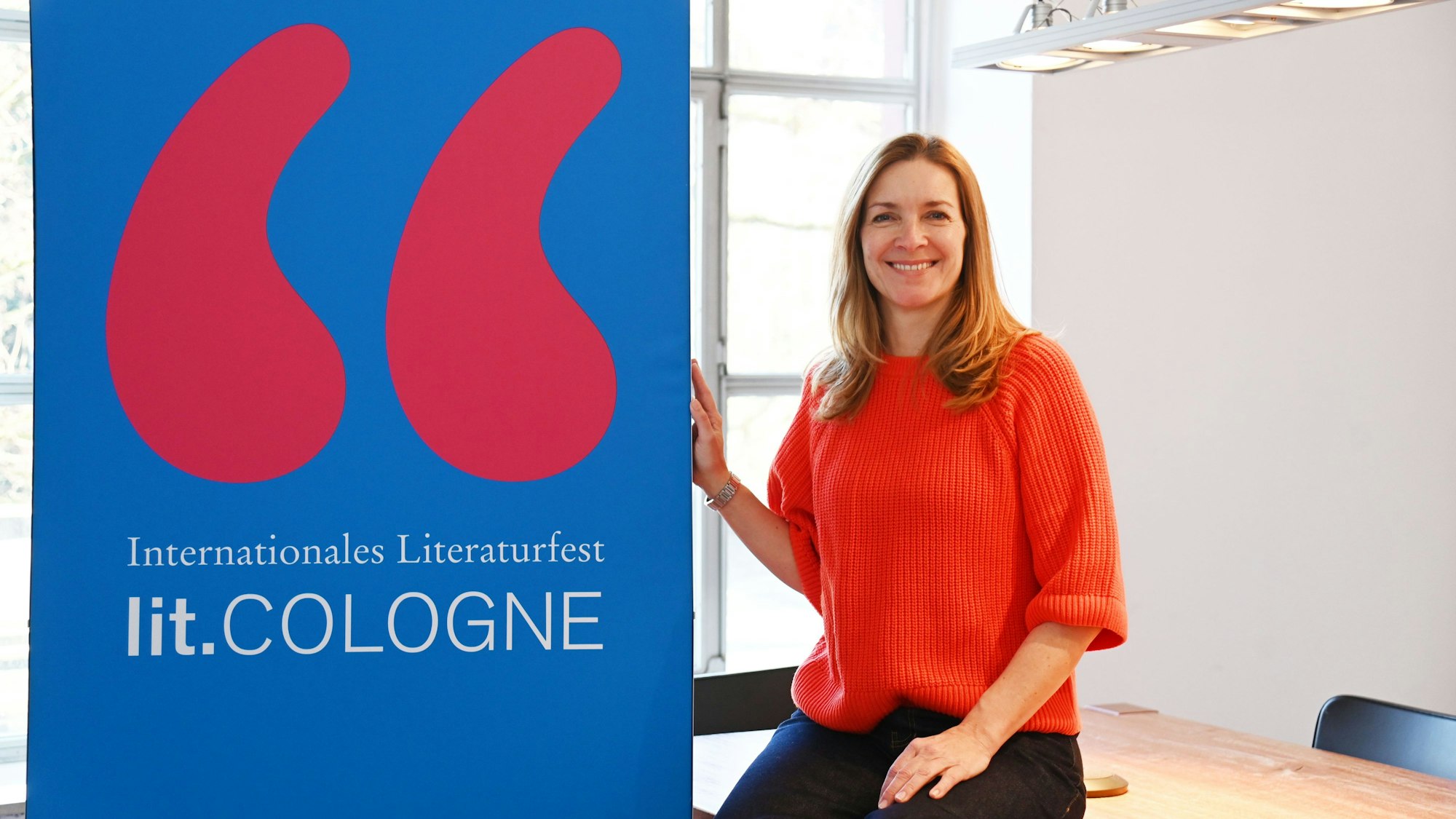 Eine Frau sitzt neben einem Schild mit der Aufschrift „Internationales Literaturfest lit.Cologne“ und lächelt in die Kamera.