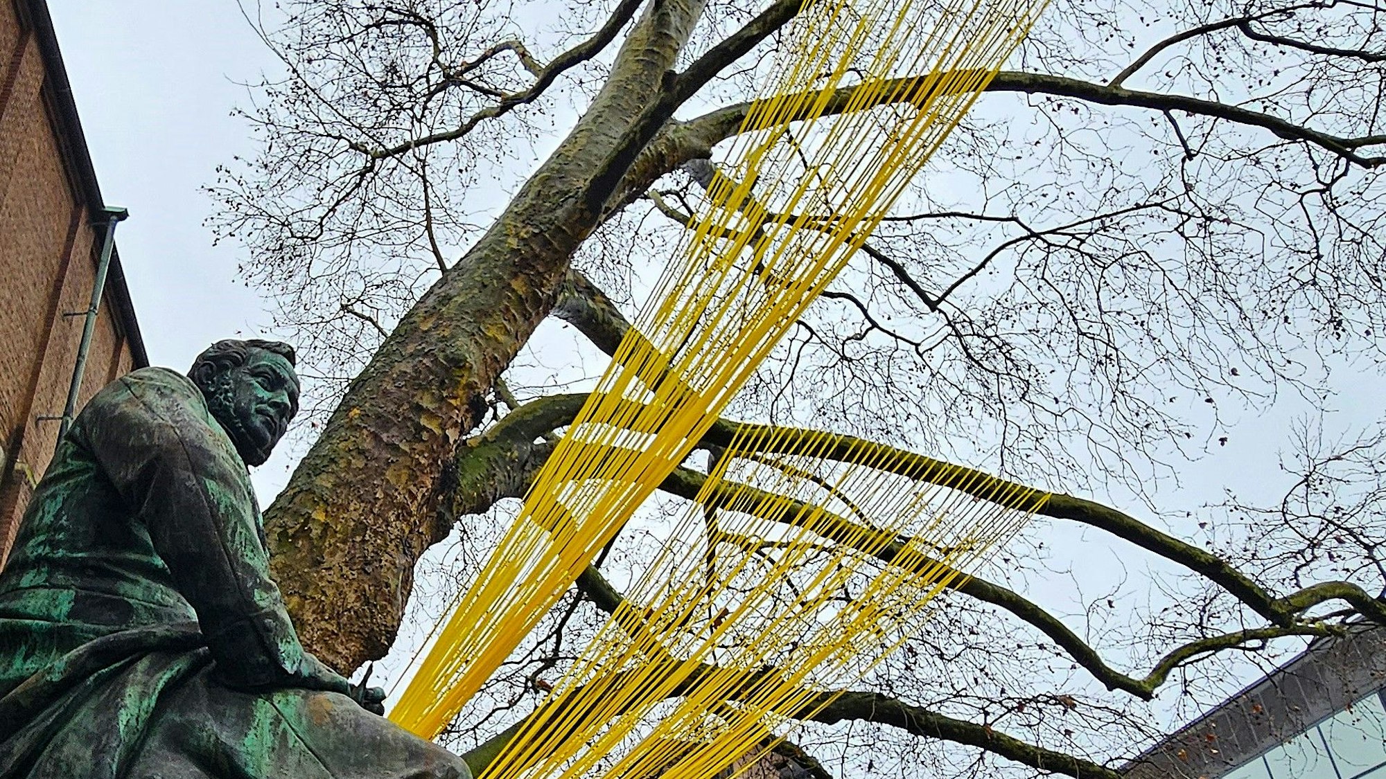 Gelbe Bänder spannen sich hinter einem Denkmal an einem Baum.