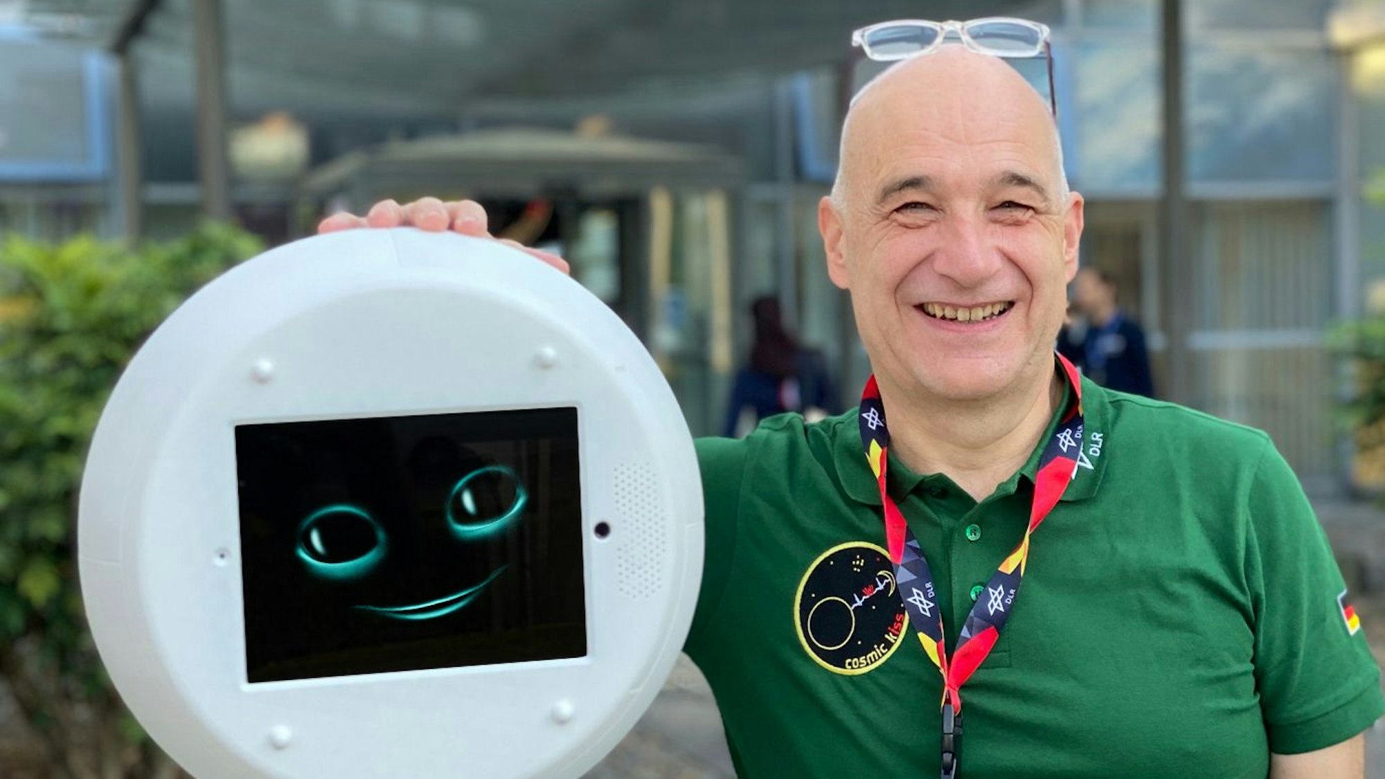Volker Schmid steht neben einem runden, medizinballrunden, flachen Roboter, der ein Gesicht mit Mund und Augen zeigt.