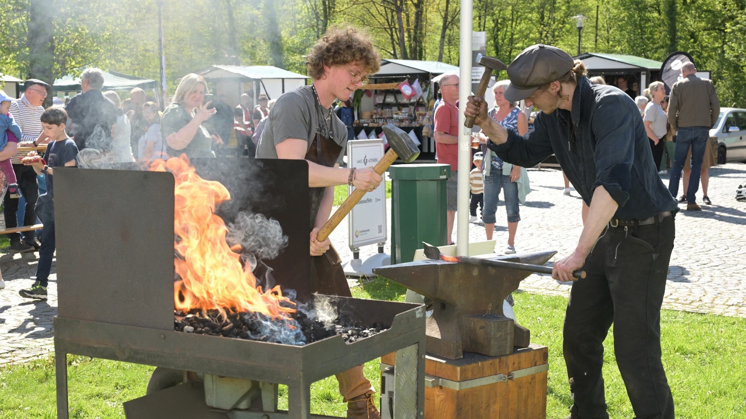 Zwei Schmiede stehen auf einer Wiese. In einer Kohleschale lodert ein Feuer. Auf dem Amboss schlagen die Männer mit dem Hammer auf glühendes Eisen ein.