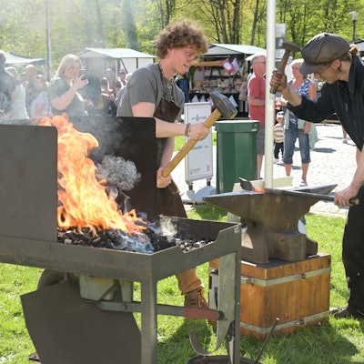 Zwei Schmiede stehen auf einer Wiese. In einer Kohleschale lodert ein Feuer. Auf dem Amboss schlagen die Männer mit dem Hammer auf glühendes Eisen ein.