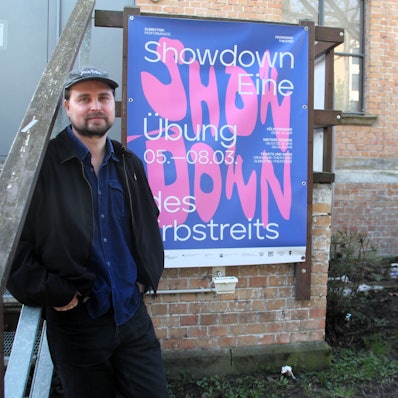Ein Mann mit Schiebermütze und Bart lehnt an einem Plakat an der Außenwand des Orangerie-Theaters im Volksgarten in Köln.