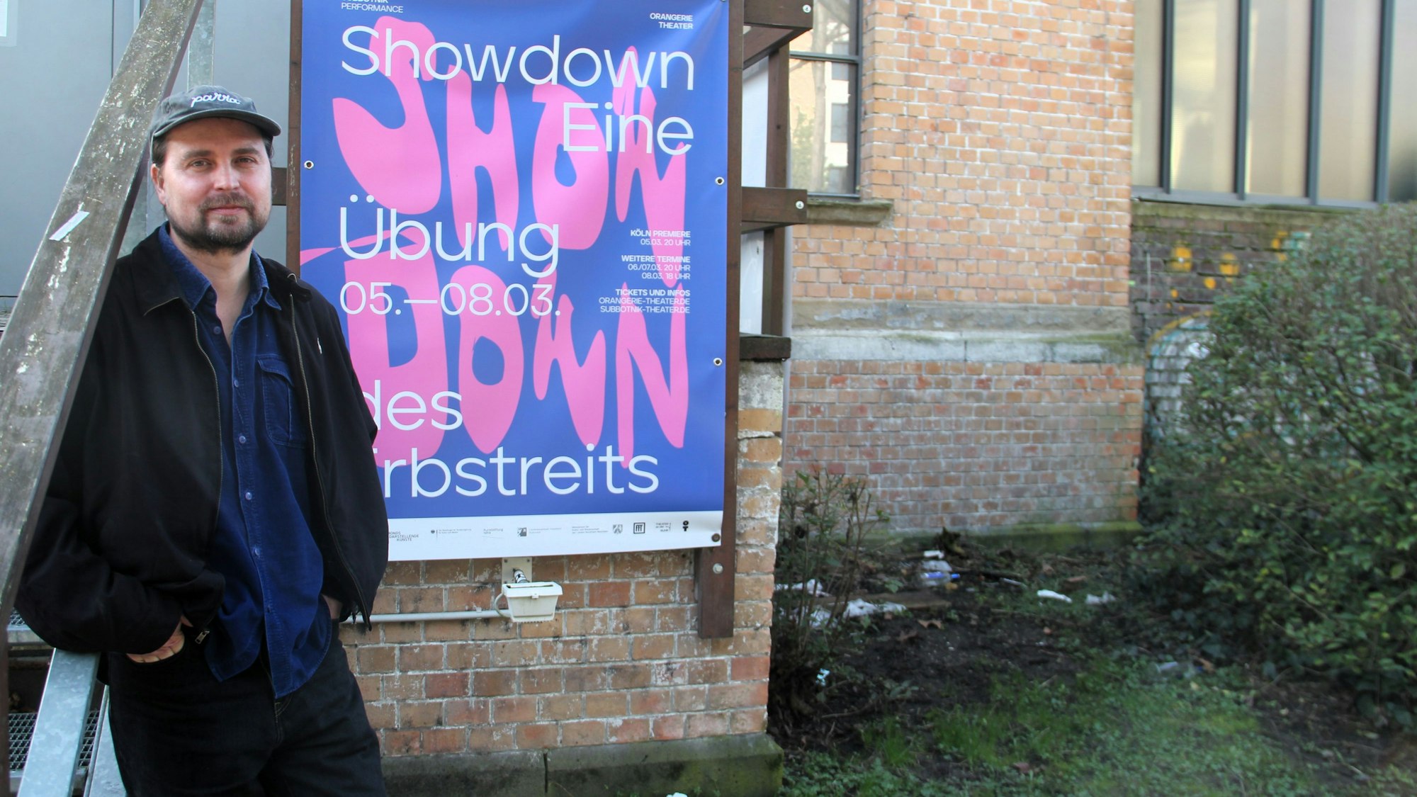 Ein Mann mit Schiebermütze und Bart lehnt an einem Plakat an der Außenwand des Orangerie-Theaters im Volksgarten in Köln.